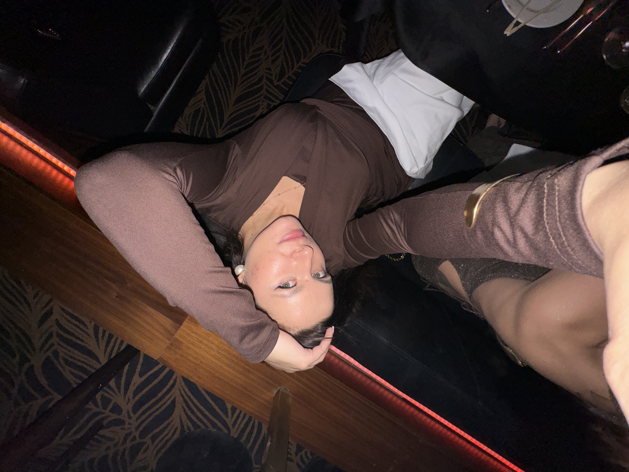 A woman in a brown long-sleeved top is lying on a dark patterned carpet next to a wooden surface with red lighting. She is wearing a white napkin or cloth under her back. A black leather booth is partially visible in the background. A table with silverware and glassware is above her, suggesting a restaurant setting. The location is likely a nightclub or upscale restaurant in Belgrade, Serbia, given the style of decor and ambiance.