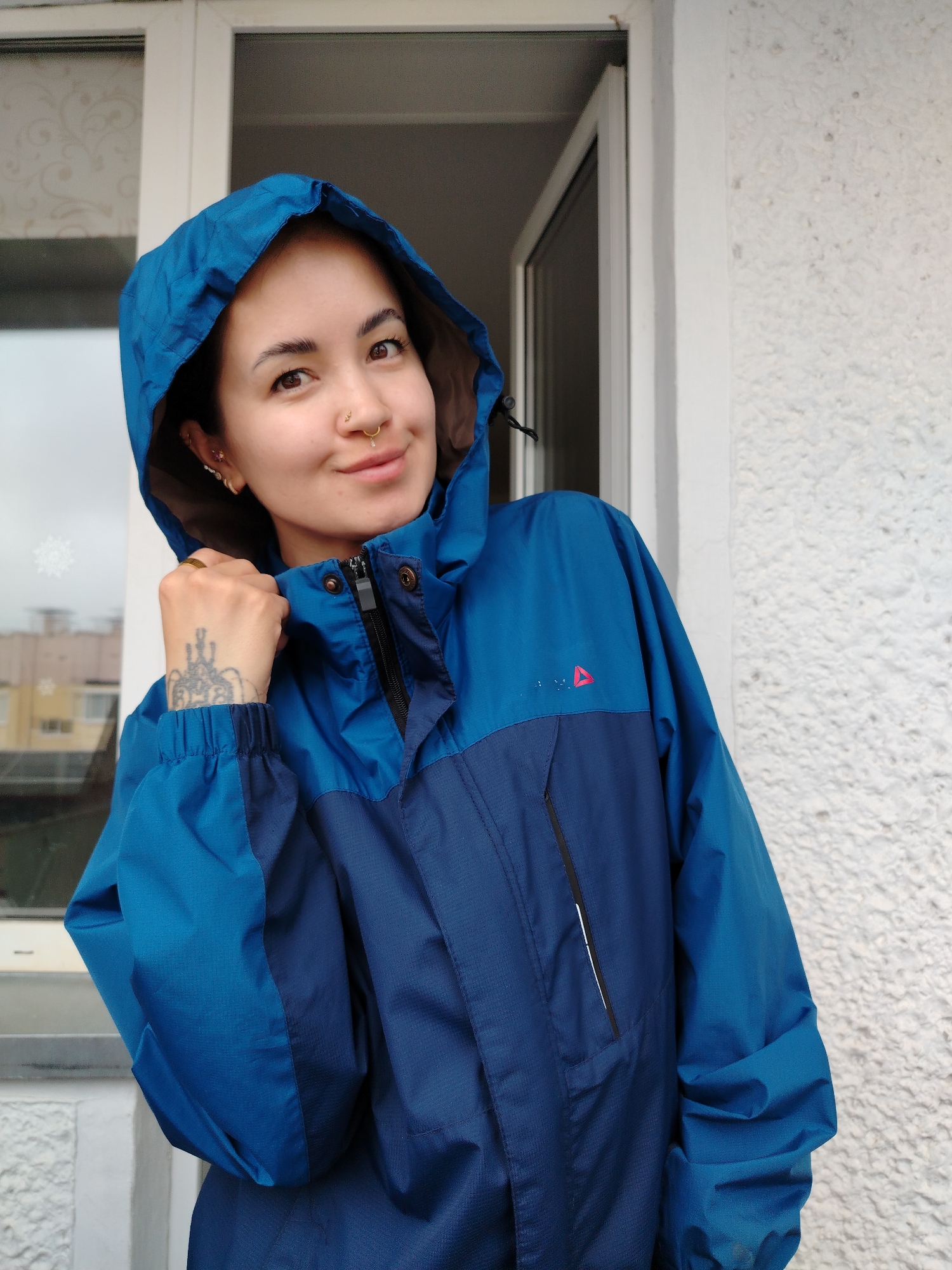 A woman with dark hair and fair skin stands in front of a white textured wall and an open doorway. She wears a blue hooded jacket with a red triangle logo on the upper right side. The jacket is zipped partway, revealing a black zipper. The hood is up, framing her face. She has multiple piercings on her left ear and a nose ring. Her left hand is raised near her neck, displaying a ring and a tattoo on her wrist. In the background, a window is visible, and beyond it, a cityscape with buildings and cloudy skies, which suggests the location is in the city of Noviy, Russia. The woman is looking towards the viewer with a slight smile.