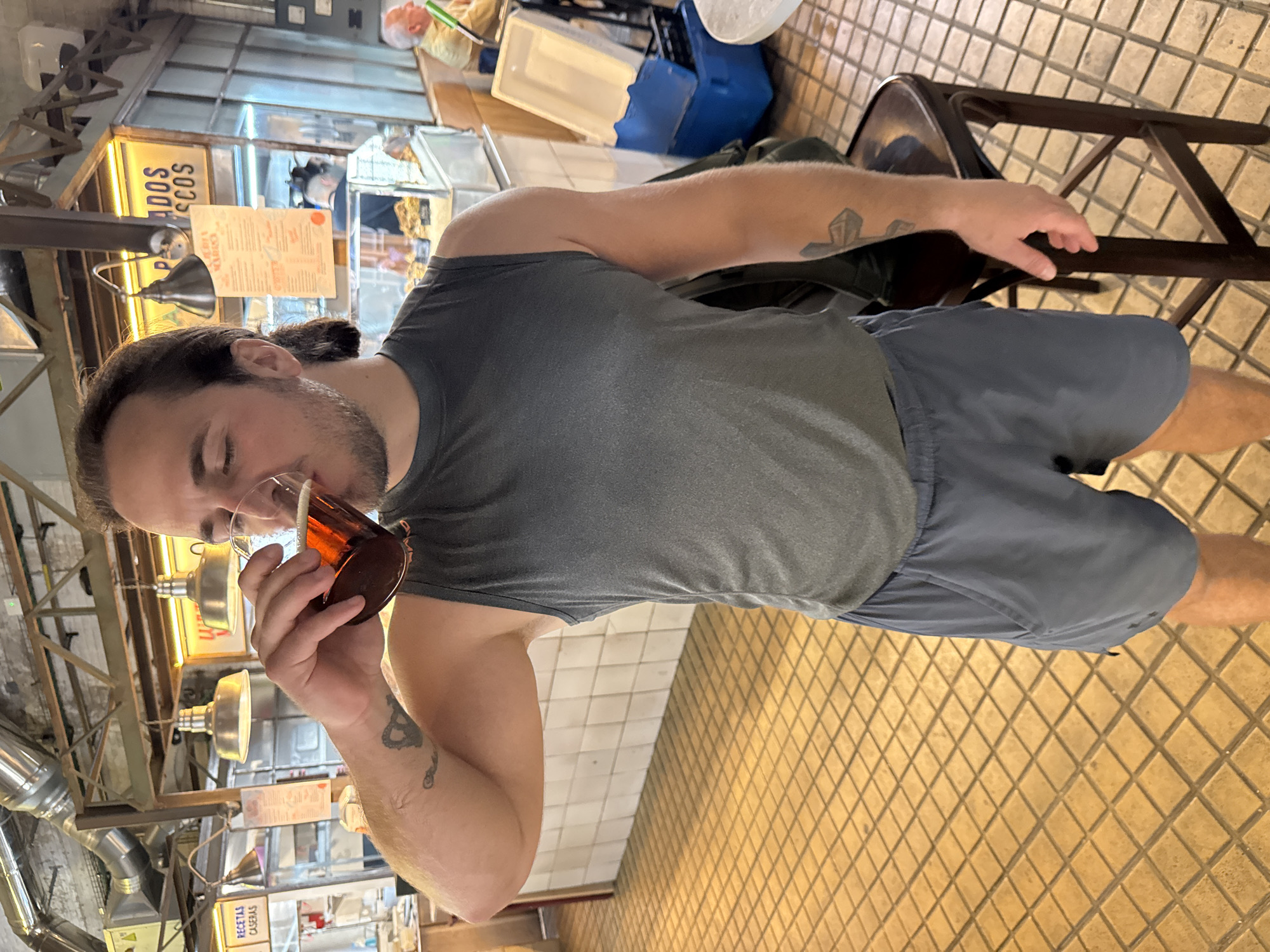 This image depicts a man enjoying a drink in what appears to be an indoor food market or casual eatery, likely in Valencia, Spain. The man, with dark hair pulled back and a beard, is wearing a grey sleeveless shirt and dark blue shorts. He has tattoos on both arms, one appearing to be a cross-like design, and another an abstract or serpentine pattern. He holds a stemmed glass filled with a dark amber liquid, possibly a beer or spirit, and is taking a sip with his eyes closed, conveying a sense of enjoyment.
The background shows elements of a bustling market environment. There are various stalls with stainless steel counters, display cases, and industrial-style pendant lamps providing illumination. The floor is covered in light brown square tiles arranged in a diamond pattern. A dark brown chair is visible next to the man. Visible text on signs includes "ADOS COS" (possibly part of a word like "pescados secos" or dried fish) and "RECETAS CASERAS" (homemade recipes), indicating food offerings. The lighting suggests it is daytime, and the overall atmosphere is relaxed and informal.