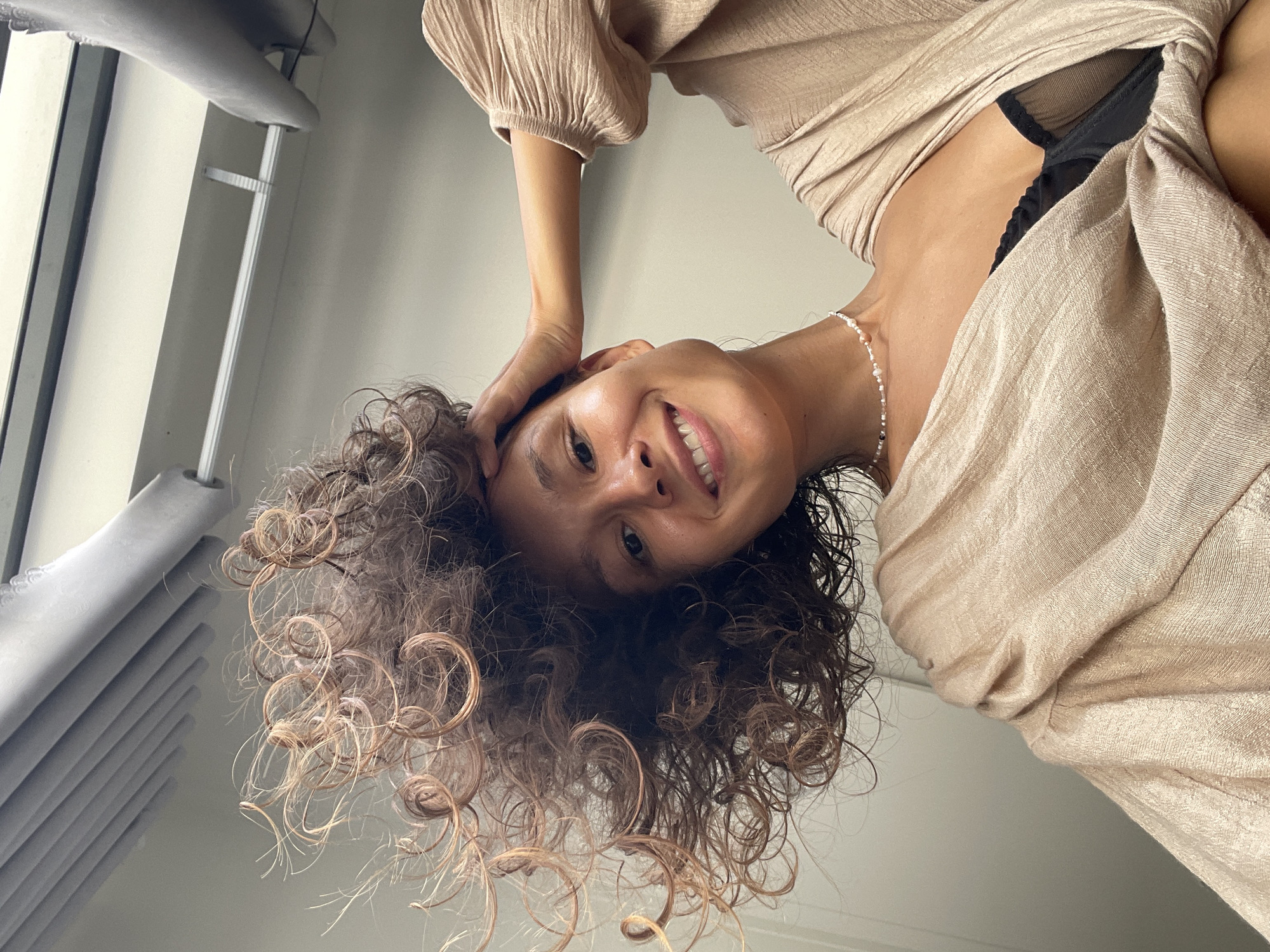 An overhead shot captures a smiling woman with curly, damp-looking brown hair, likely indoors in Mũi Né, Vietnam. Her head is tilted, and she looks directly into the camera with a happy expression, one hand gently resting on the side of her head. She is wearing a light beige or gold-colored top or dress with a textured, slightly crinkled fabric and puffed sleeves. A black bra is visible underneath the sheer material of her top. Around her neck is a delicate white beaded necklace with subtle peach or pink accents.
The background consists of a light-colored wall or ceiling, with a window frame and part of a pleated blind visible on the left side of the image, suggesting a daytime setting with ambient light. No specific time of day or weather can be determined, and no other location-specific cues are present within the image itself. There is no visible text. The perspective is from below, looking up at the woman, creating an intimate and engaging portrait.