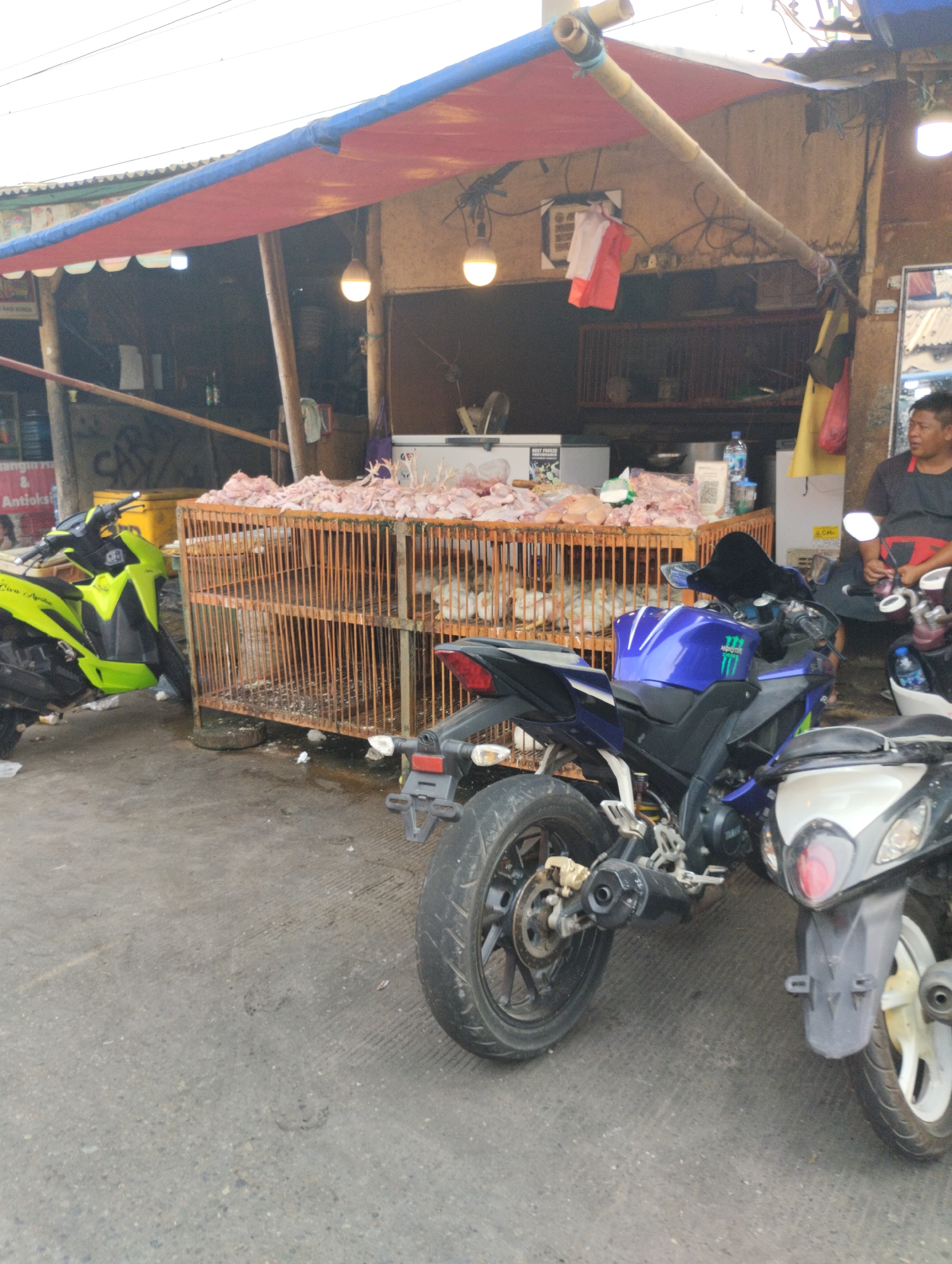 A street-level, medium shot captures an outdoor market stall in Bekasi, Indonesia, with a focus on raw poultry displayed in a large, rustic cage. The stall is partially covered by a red and blue awning, supported by bamboo poles. The cage, made of metal bars, is filled with raw chickens. In front of the cage, a bright yellow motorcycle is parked, and to the right, a blue motorcycle and a white and black motorcycle are visible. A man in a dark shirt and apron is standing behind the motorcycles, facing away from the camera. The ground is paved and appears somewhat weathered. The background shows more market stalls and structures, suggesting a bustling commercial area. Overhead, electrical wires are visible against a bright sky.