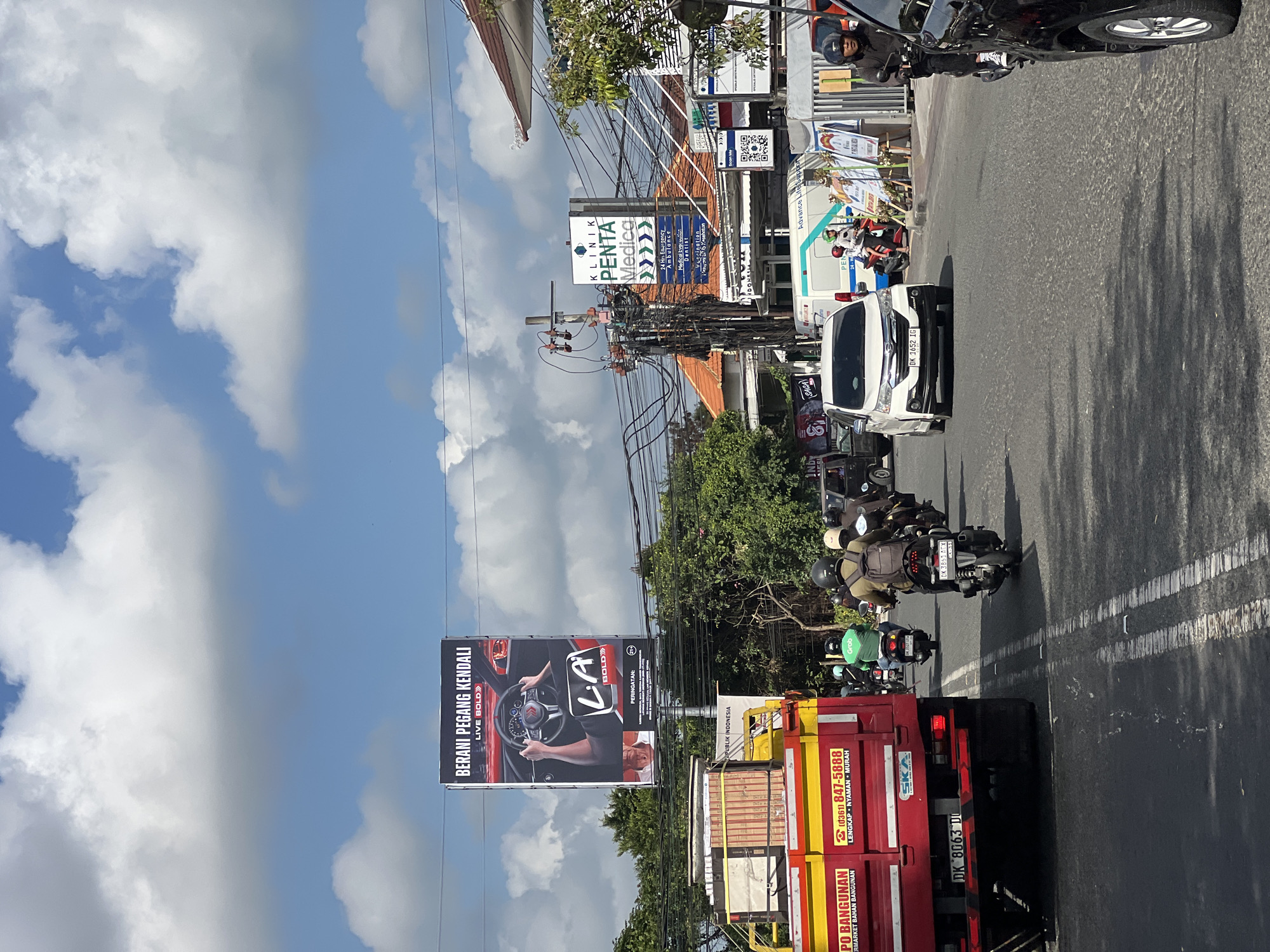 The image captures a street scene in Denpasar, Indonesia, on a bright, cloudy day. In the foreground, a red truck with a yellow cargo bed is parked, its side displaying the text "IPO BANGUNAN" and a phone number. Several motorcycles and a white car are navigating the road. A prominent billboard for "LA Bold" cigarettes is visible overhead, featuring an image of hands on a steering wheel. To the right, a sign for "KLINIK PENTA Medica" indicates various medical services offered, including a 24-hour emergency ambulance. The environment is characterized by a busy street with overhead power lines, trees, and buildings lining the sides of the road. The sky is a vibrant blue, dotted with white clouds.