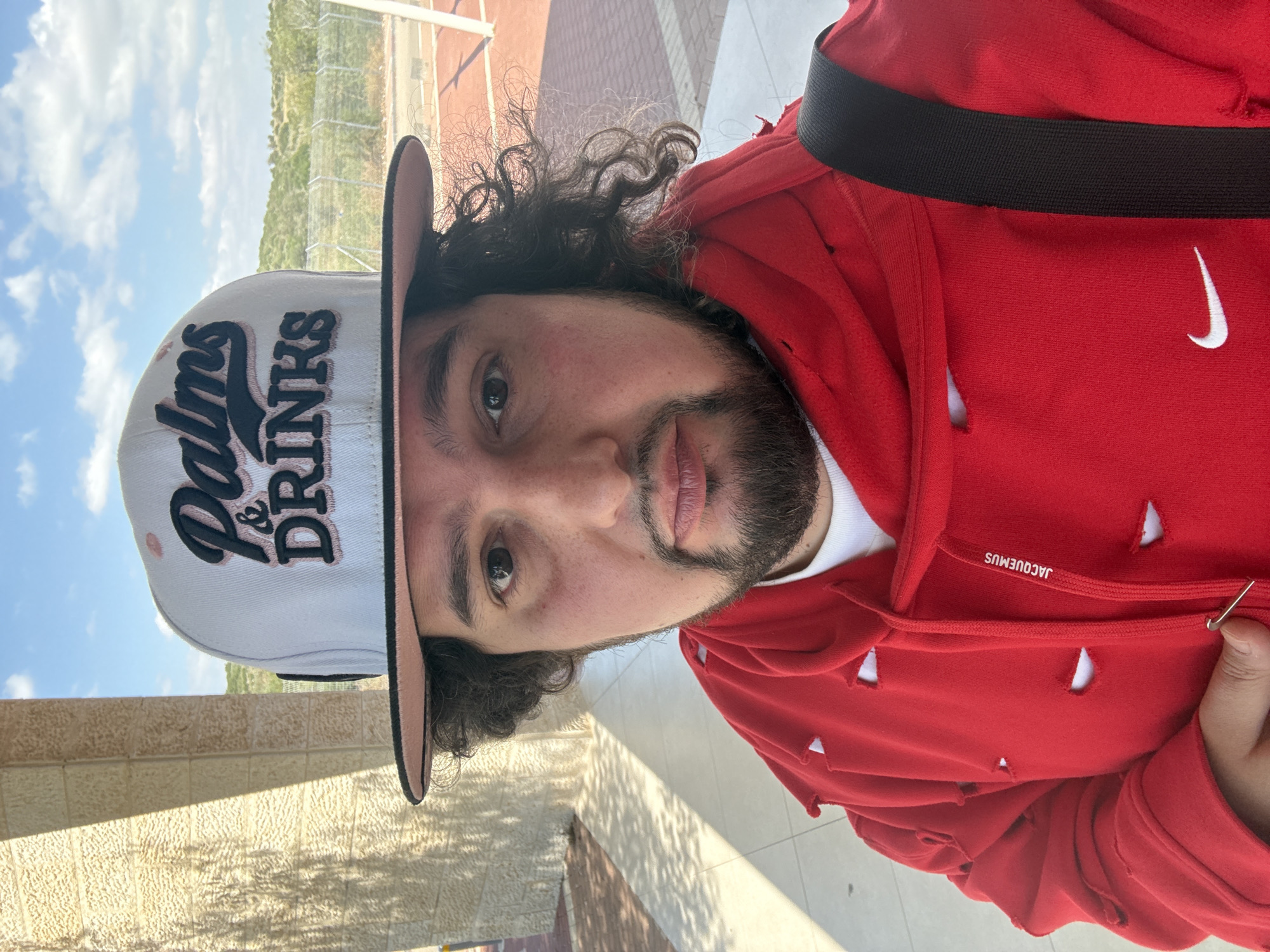 A man is pictured from the chest up, wearing a red Nike hooded sweatshirt with a black strap across his shoulder, and a light gray hat with the words "Palms & Drinks" embroidered in black. The hoodie has several small, crescent-shaped cutouts and the brand name "JACQUEMUS" printed on the collar. The man has curly dark hair and a beard. The background includes a tiled walkway, a low stone wall, and a view of trees and a running track under a partly cloudy sky. The location is Modiin-Maccabim-Reut, Israel. A safety pin is visible, held by his right hand, pinning part of the hoodie.