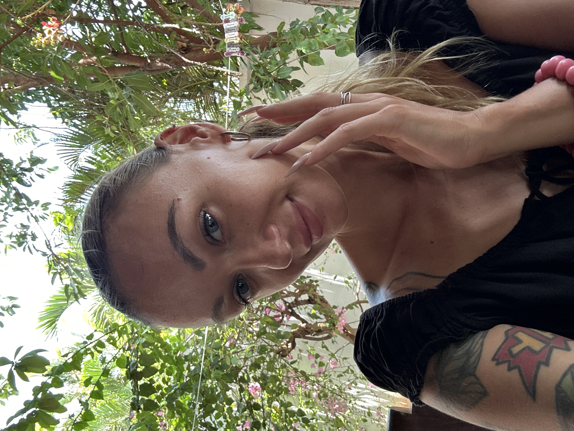 A woman is positioned in the foreground, with a tilt upwards revealing foliage in the background. She has blond hair and light skin, with visible tattoos on her chest and arm. Her hand is raised near her face, adorned with rings. The backdrop includes tropical vegetation, suggesting an outdoor setting. There are no other people present. It is hard to pinpoint location due to foliage.