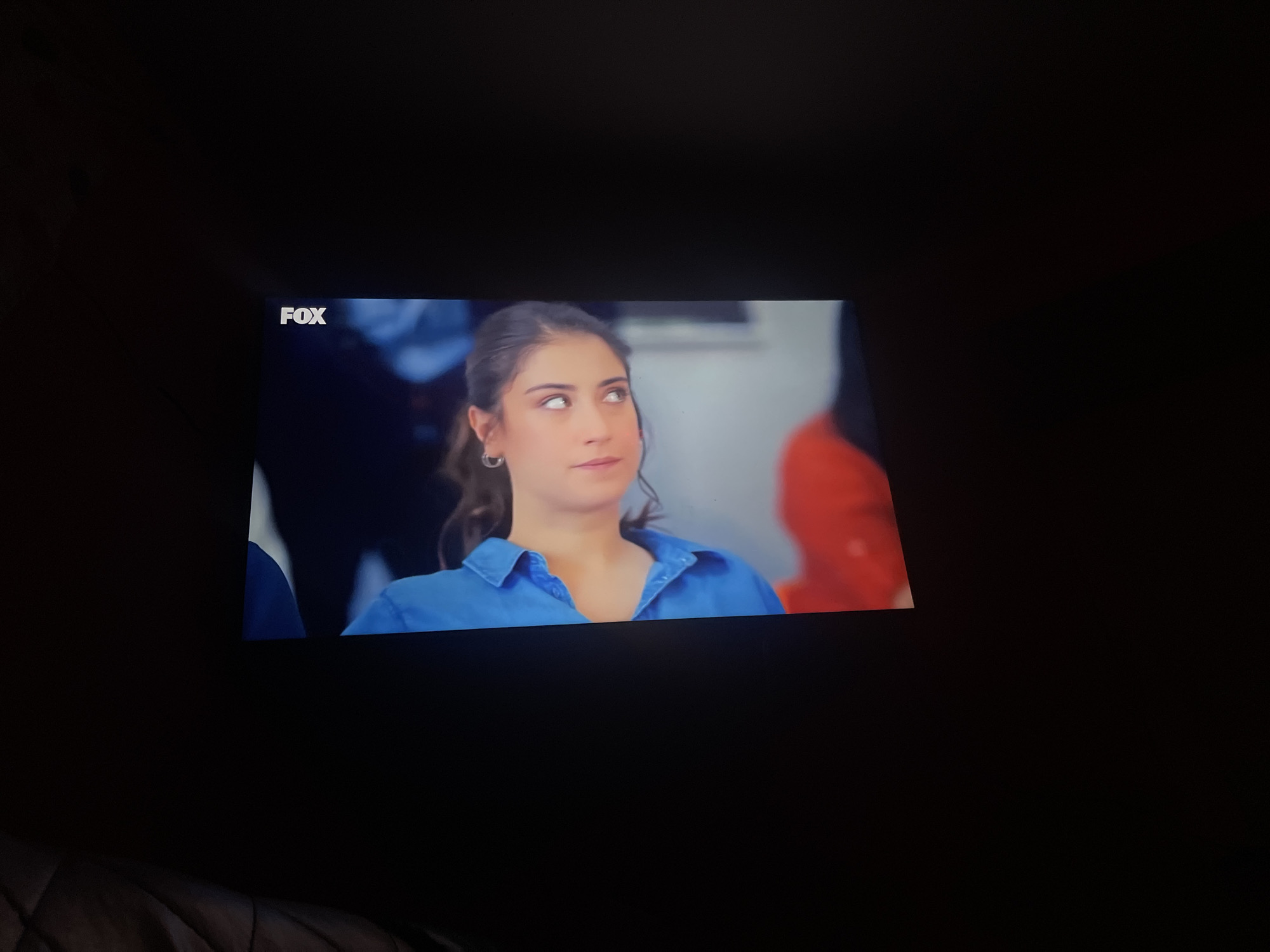 The image shows a television screen displaying a scene from a show. A young woman with brown hair pulled back is looking upwards with her eyes rolled back. She is wearing a blue collared shirt. The "FOX" logo is visible in the upper left corner of the screen. The background is blurred, with some hints of other people and a red object on the right side. The lower portion of the image shows the dark, textured surface of furniture, likely a couch, and the screen's frame. The scene is not identifiable as being in Dolni Dabnik, Bulgaria.