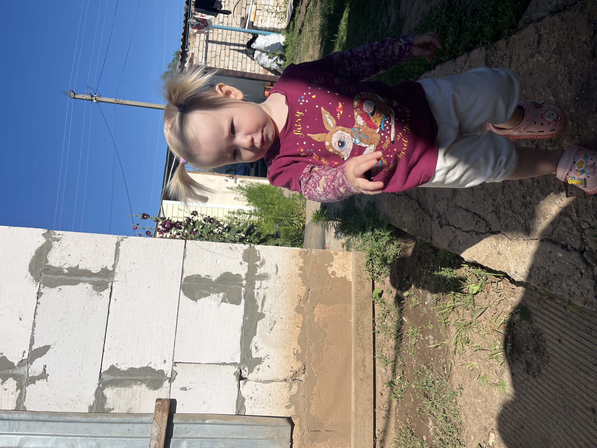 A blonde-haired toddler with pigtails walks on a concrete path in what appears to be a rural or suburban area. The toddler is wearing a burgundy long-sleeved shirt with a deer graphic and the words "Fairy" and "Secret Forest" on it, white pants, and pink Crocs. The toddler is looking towards the right of the frame with a serious expression.
In the background, to the left, is a partially constructed white block wall. To the right, there is a brick building, a clothesline with laundry hanging, and a wooden fence. The ground is covered with dirt, grass, and some gravel. The sky is clear and blue. Shadows are cast on the path, indicating a sunny day.
