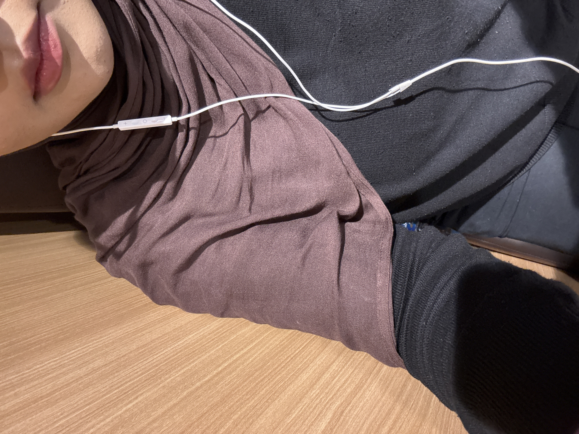 The image is a close-up shot of a person wearing a dark brown hijab and a black long-sleeved shirt, with white earphones visible. The background appears to be a light brown wooden surface, possibly a wall or furniture. The person's lips are partially visible, painted with a subtle pink lipstick. The lighting suggests an indoor setting. The image is cropped to focus on the upper body and face, excluding the lower body and the full surroundings. There is no identifiable location within West Jakarta, Indonesia, visible in the image.