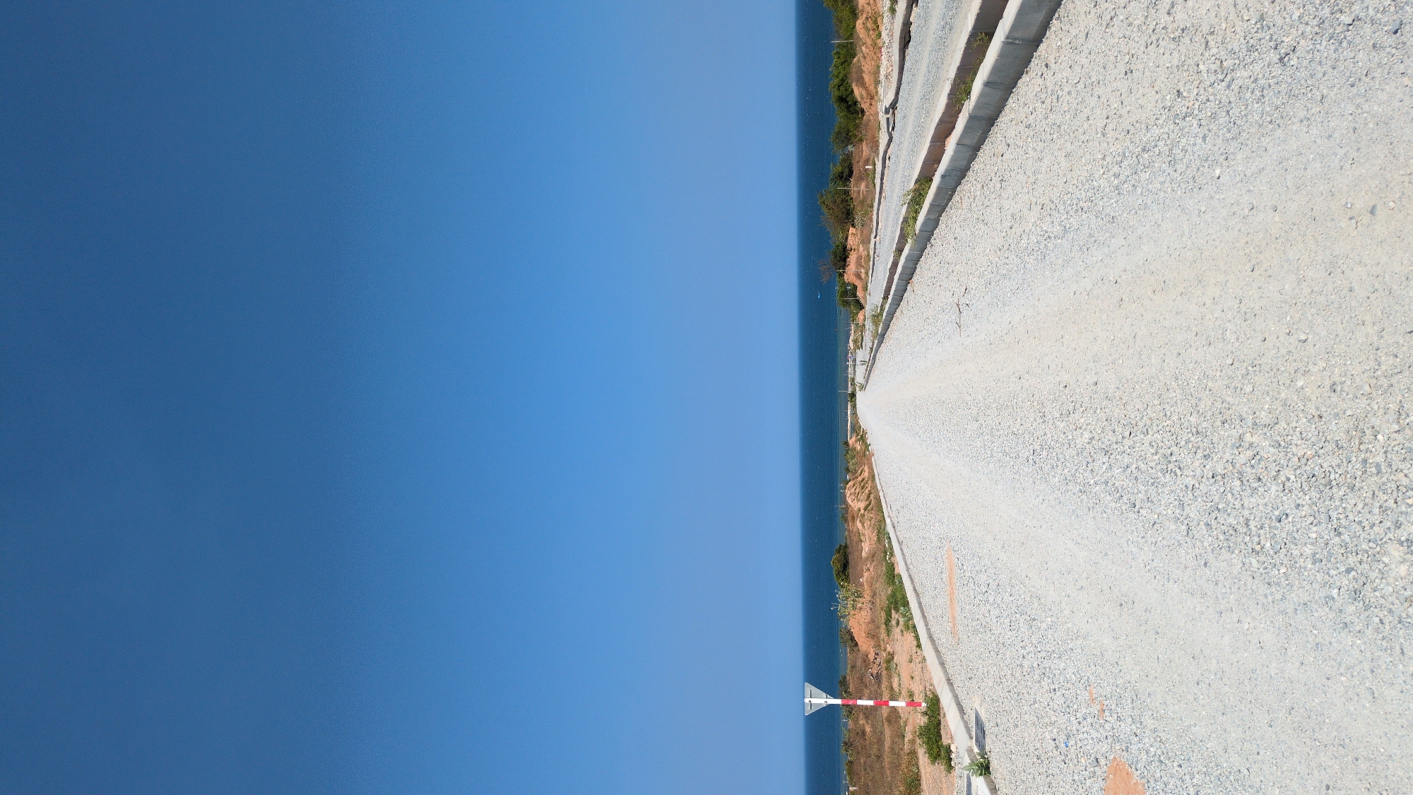 The image depicts a road in Phuoc Dinh, Vietnam. The road surface is gravel, and it runs parallel to the coastline. The sea is visible on the left side of the road, meeting the clear blue sky at the horizon. A red and white striped pole stands near the road, likely marking a boundary or hazard. Vegetation and some built infrastructure are present between the road and the sea.