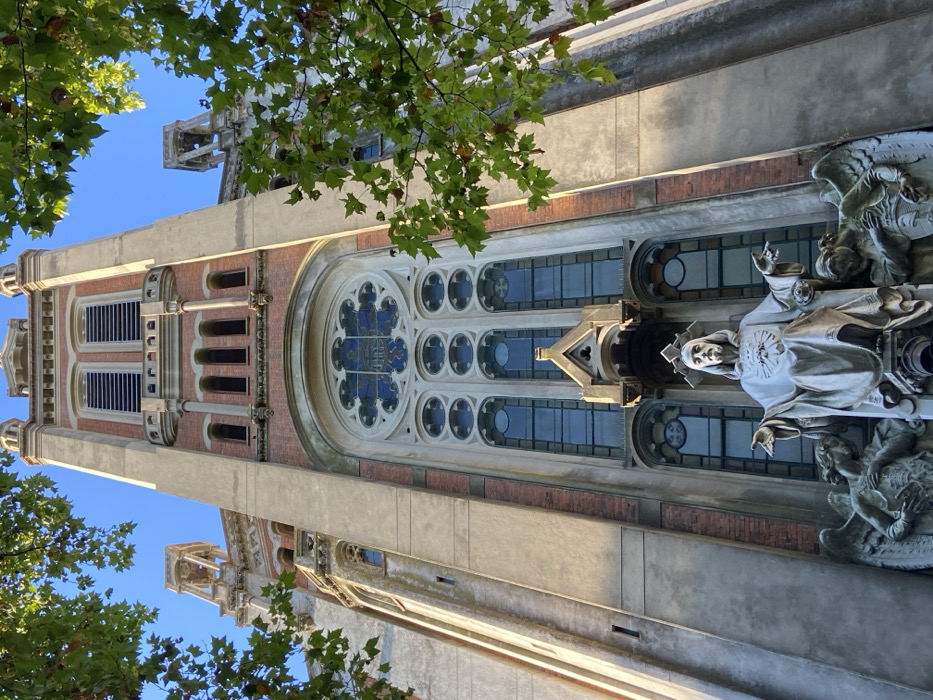 A tall church tower dominates the frame, partially obscured by tree branches in the upper left and lower left. The tower's architecture features multiple windows, including a large, round one with blue accents. To the right of the windows stands a statue with its arms outstretched, framed by angels. Sunlight casts shadows across the building's textured exterior.