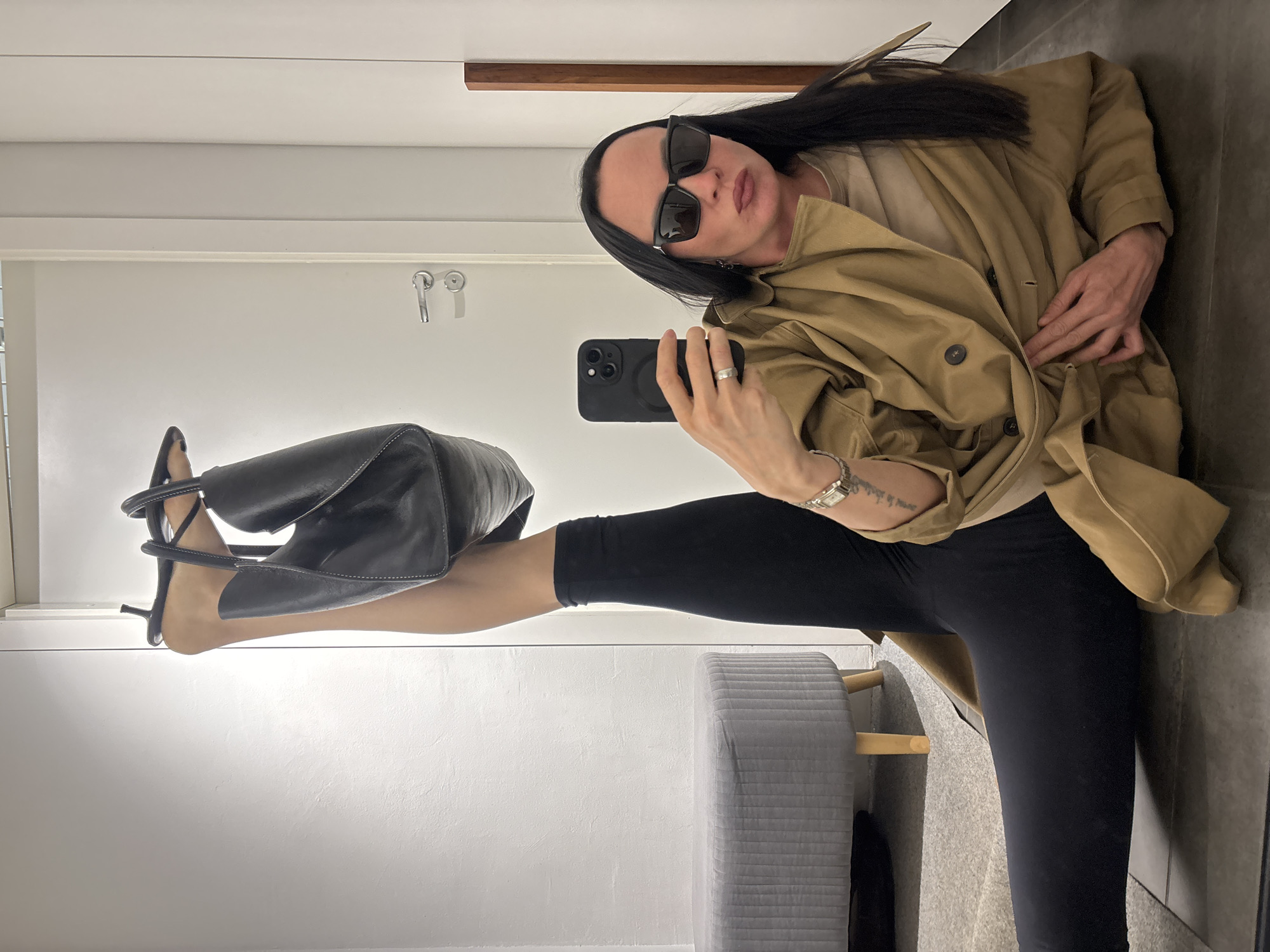 A person is taking a selfie in a room with a white door and grey walls. The person is wearing sunglasses, a light brown trench coat, and black leggings. They are holding a black smartphone in their right hand. A black leather handbag is suspended by the person's left foot, which is extended upwards. A grey upholstered stool with light wood legs is visible near the person's legs. There is a coat hook on the door. A horizontal brown strip is on the wall near the ceiling. The location, Chaikovsky, Russia, is not visible.