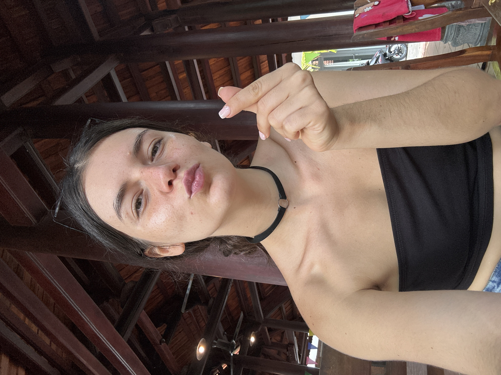 A young woman, with freckles across her nose and cheeks, is pictured in a close-up, chest-up shot. She has dark hair pulled back and is wearing a black strapless top and a black choker necklace. Her lips are pursed, and her right hand is held up, forming a heart shape with her thumb and index finger. The background reveals an open-air structure with dark wooden beams and pillars, suggesting a covered outdoor area. Lighting fixtures are visible on the ceiling. To the right, a blurred glimpse of a motorcycle and red fabric draped over a chair can be seen, indicating an outdoor setting, possibly a cafe or restaurant, in Mũi Né, Vietnam.