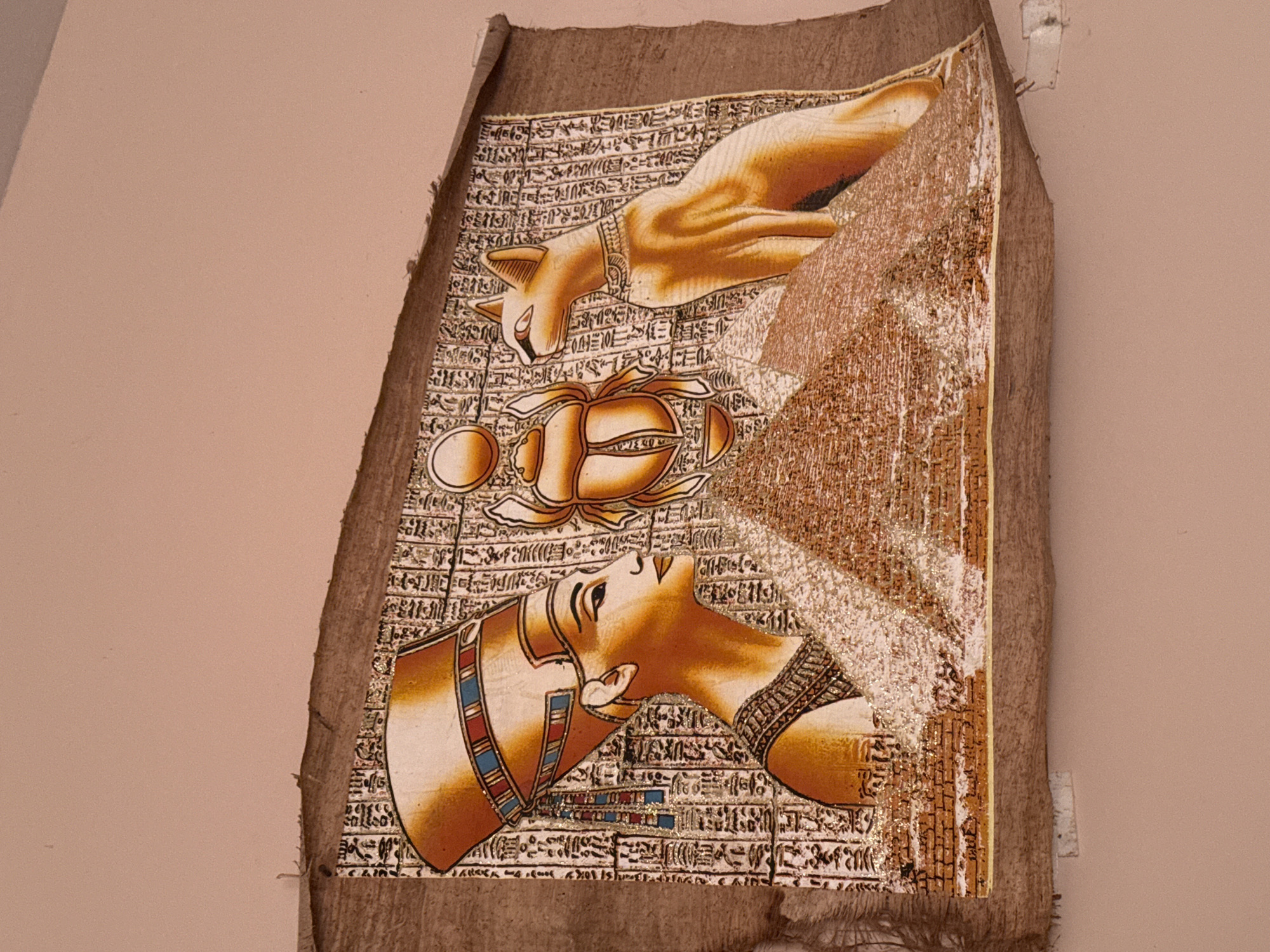 The image depicts a framed papyrus painting depicting Egyptian motifs. On the left, a profile portrait of Queen Nefertiti is shown wearing her distinctive crown, adorned with stripes of blue, red, and gold. She is rendered in warm golden hues. To her right, a scarab beetle with outstretched wings is centrally positioned above a stylized representation of a pyramid, also rendered in golden tones and textured to resemble brickwork. To the right of the scarab and pyramid, a seated cat with alert ears and a jeweled collar is depicted in a golden hue. The background of the painting is filled with hieroglyphic inscriptions, and metallic gold accents are visible throughout the artwork, particularly on the pyramids and surrounding hieroglyphs. The papyrus itself appears aged and has a rough, textured surface with frayed edges. The painting is affixed to a plain, light pink wall, which suggests it is displayed indoors. The image does not provide any visual cues that would indicate its location in Montana, Bulgaria.