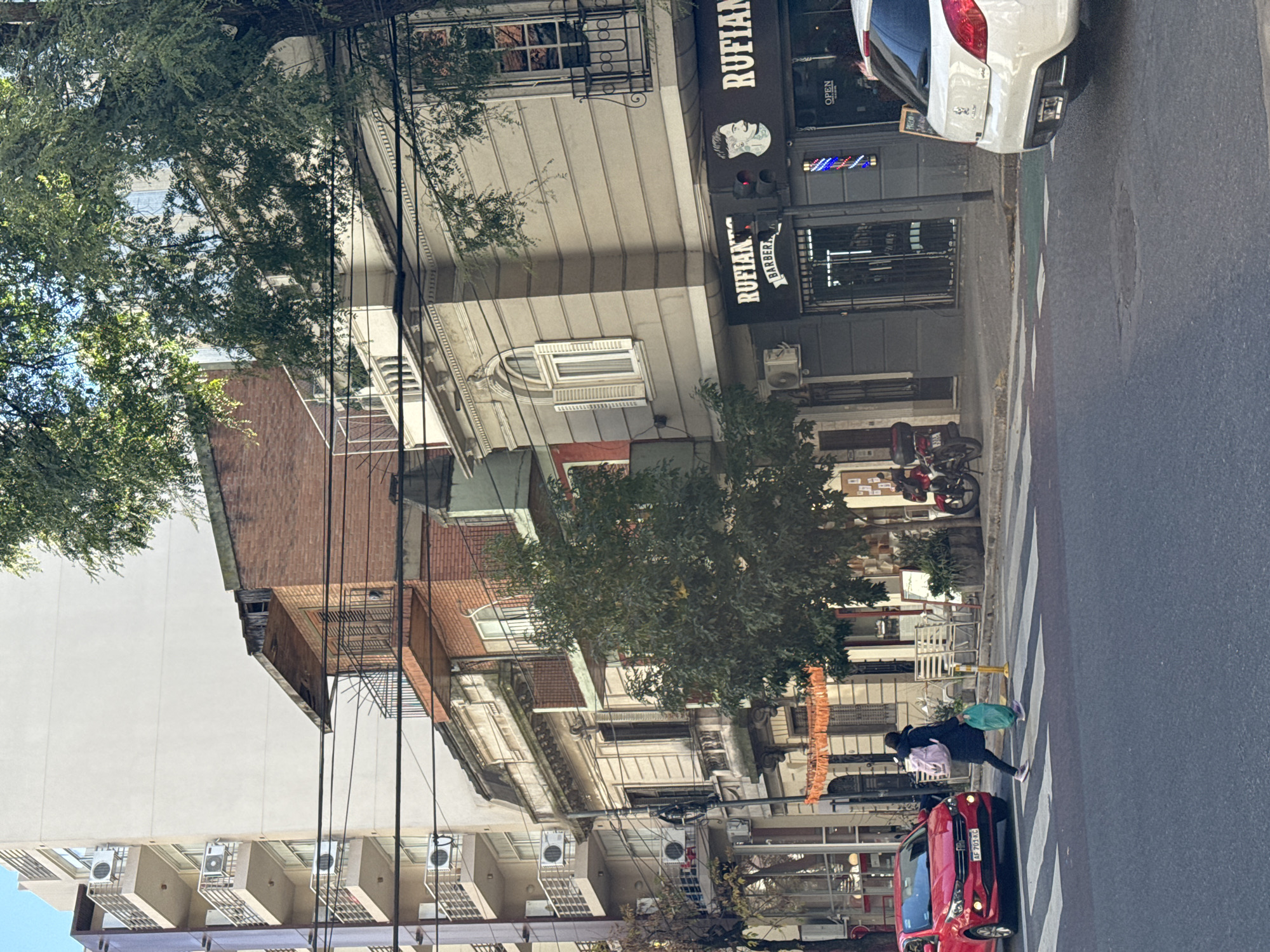 A street scene in Buenos Aires, Argentina, shows a pedestrian crossing the street at a crosswalk. The pedestrian, dressed in dark clothing and carrying bags, is walking from left to right. A red car is stopped at the crosswalk on the left. A white car is parked at the curb on the right in front of a barber shop called "RUFIAN." The barber shop has a traditional barber pole symbol. The architecture includes a mix of building styles and heights, with trees lining the street. Overhead, a network of utility wires is visible.