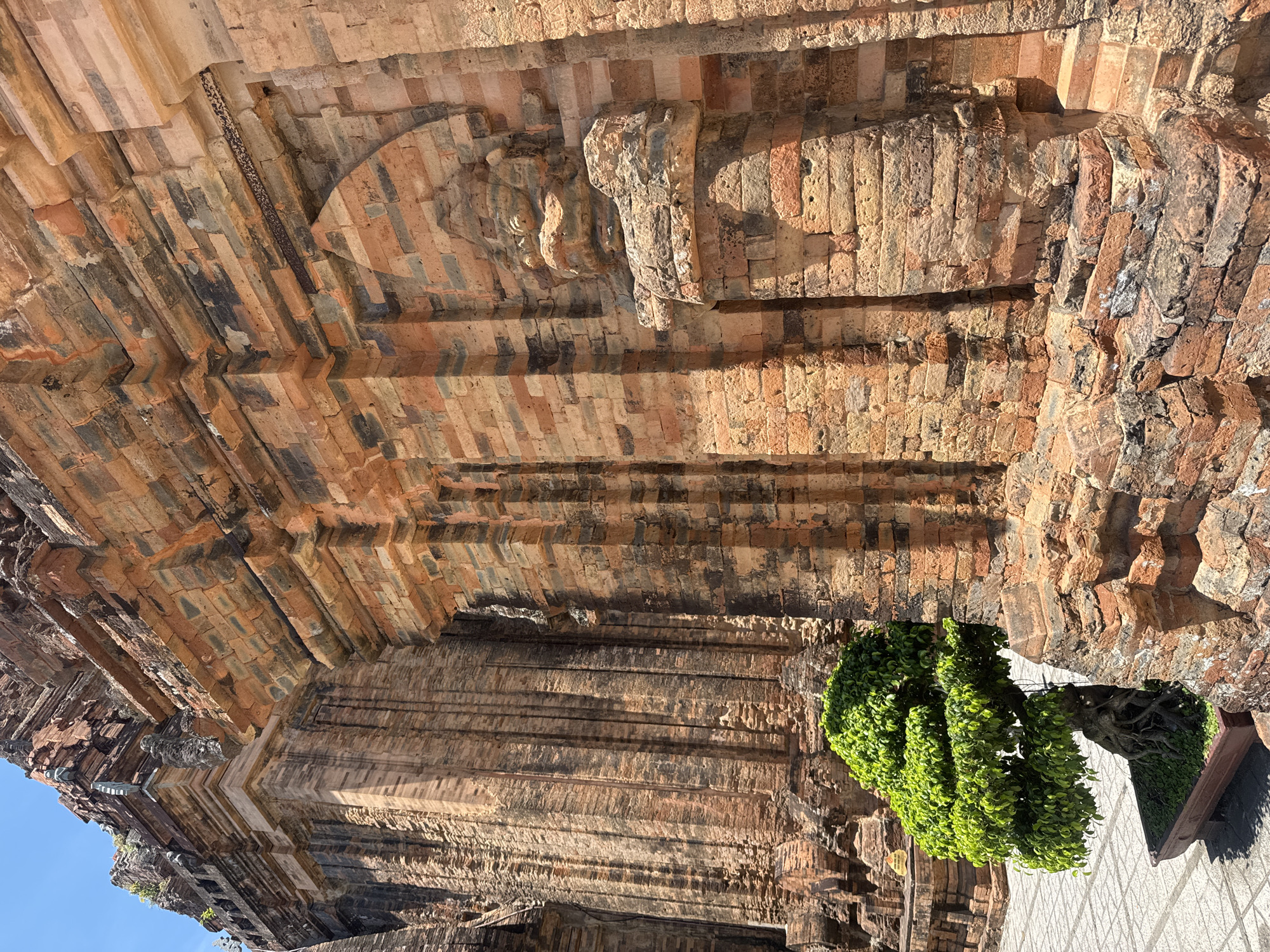The image shows the Cham Towers in Nha Trang, Vietnam, a historical religious site made of red bricks. The towers have intricate carvings and architectural details, showing signs of age and weathering. In the foreground, a meticulously shaped bonsai tree in a rectangular planter adds a touch of vibrant green against the warm tones of the brickwork. The ground around the towers is paved with patterned tiles. The sky above is a clear blue.