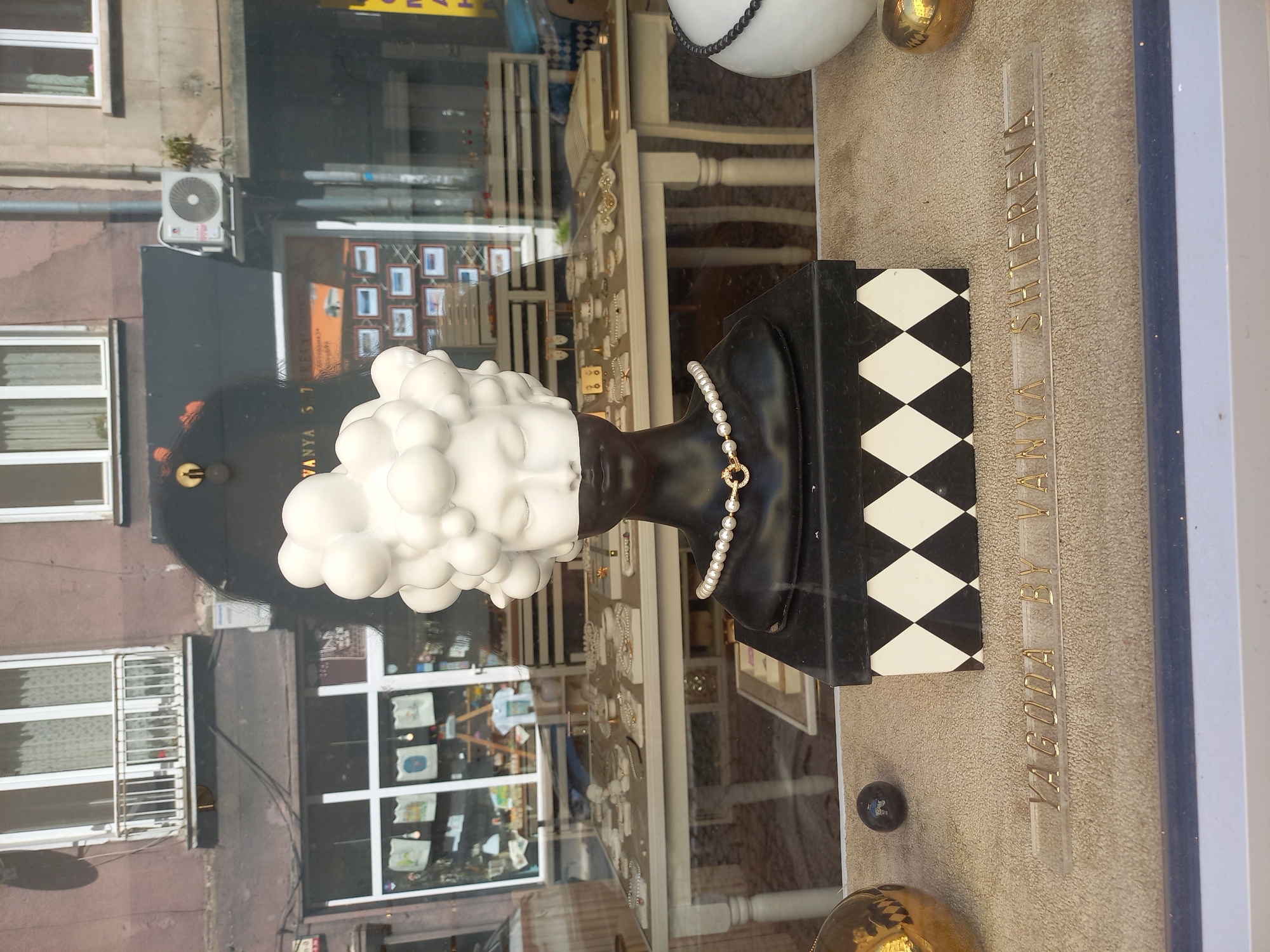 This image captures a display window in Sofia, Bulgaria, featuring an artistic bust of a woman with a hairstyle made of white spheres. The bust is black from the neck down, adorned with a pearl necklace featuring a gold clasp. It sits atop a black and white diamond-patterned box. In the foreground, a clear acrylic strip displays the gold lettering "YAGODA BY VANYA SHTEREVA". To the left of the bust, a large golden sphere and a small black sphere are visible. The background of the window shows a street view with shops and buildings, including an air conditioning unit mounted on a wall and a balcony with a white railing. The overall scene suggests a retail display for jewelry or fashion accessories.