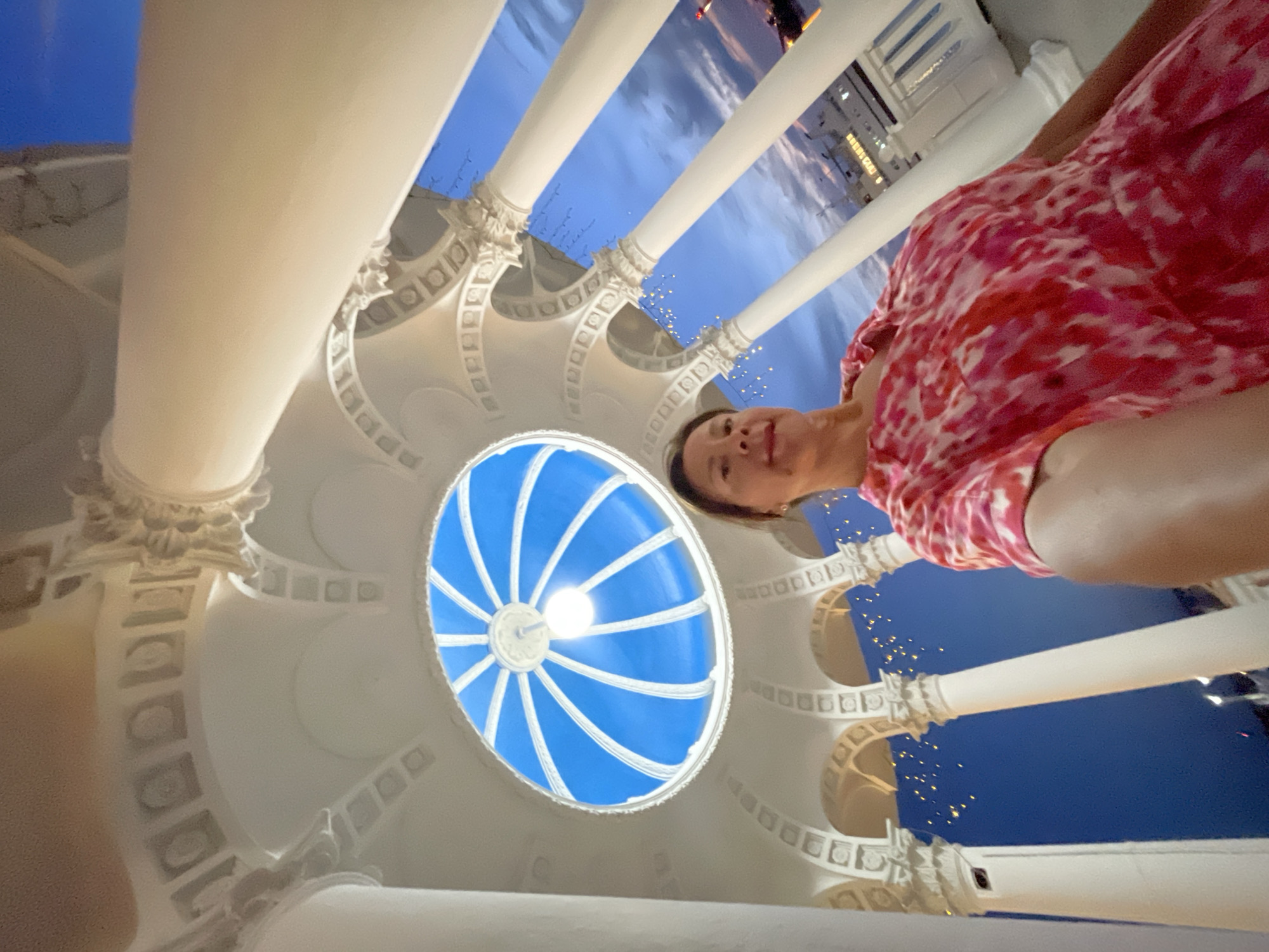 An outdoor selfie is taken from a low angle, looking up at a woman and an ornate white structure with a blue dome overhead. The woman, wearing a pink and red tie-dye dress, is in the foreground, her arm extended to capture the shot. The structure features classical columns and intricate detailing, with small lights strung along its upper portions. Beyond the structure, the sky is a deep blue, transitioning to twilight hues, with a large ship visible in the distance. The scene appears to be set at dusk or early evening. The building with the blue dome and columns is likely part of the Sochi Dendrarium's "Italy Garden" or a similar ornamental area within the arboretum complex, a popular tourist attraction in Sochi, Russia.