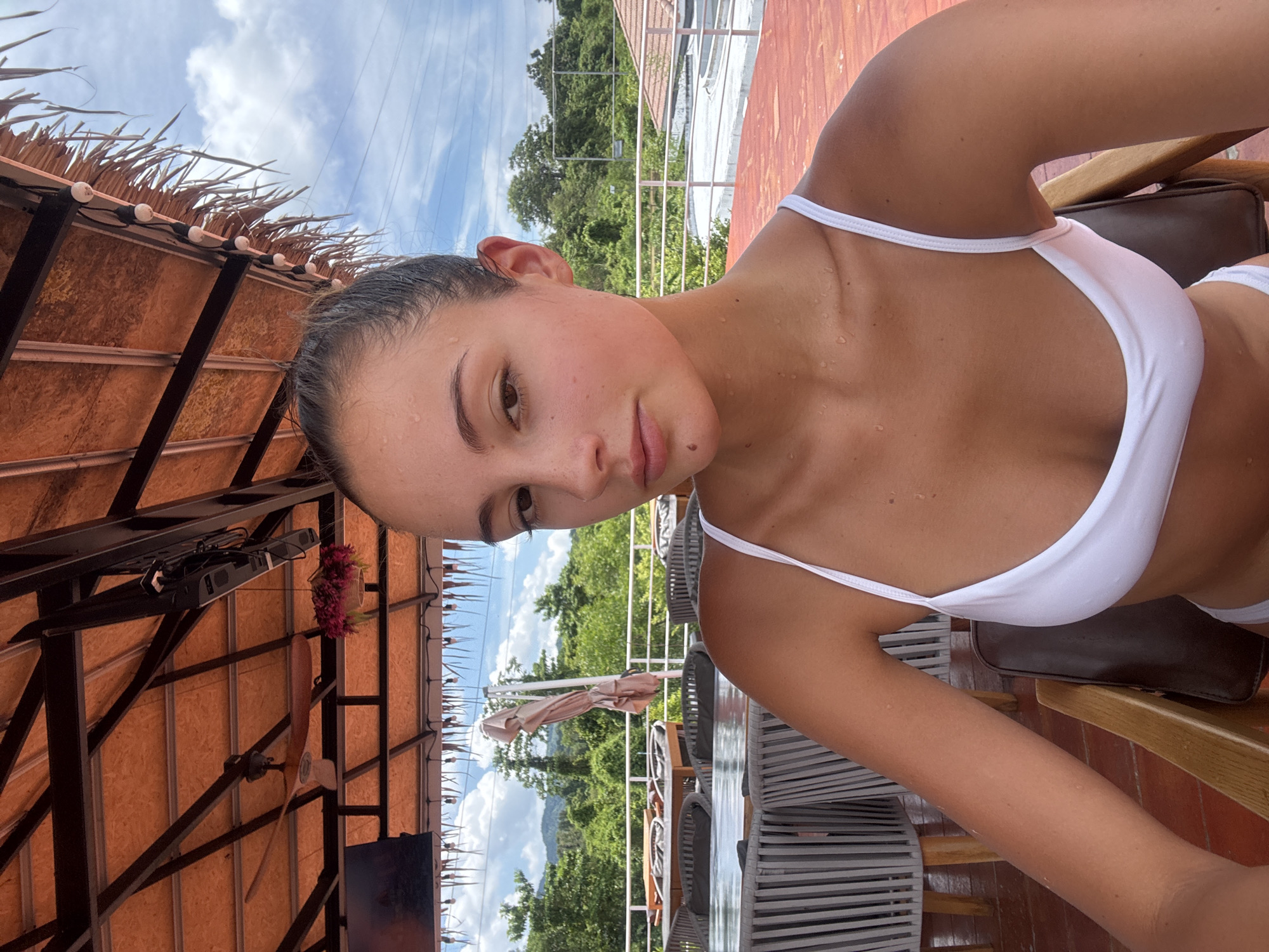 The image depicts a woman with fair skin and brown hair, wearing a white bikini top, seated outdoors. The setting appears to be a restaurant or lounge in Kathu, Thailand. The background includes lush green trees and mountains, suggesting a tropical location. The woman is positioned under a thatched roof structure with lights, a ceiling fan, and a television. The tables and chairs are visible, indicating an outdoor dining or relaxation area. She has droplets of water on her face and chest, possibly from humidity or swimming.