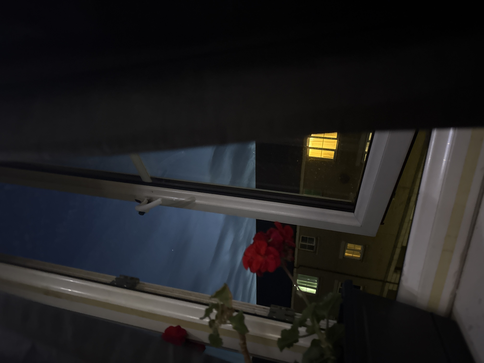 A nighttime view looking out from a window partially open, likely in Great Yarmouth, United Kingdom, as it depicts a common residential scene typical of the area. The open window reveals the night sky and glimpses of a building across from the window, suggesting a residential neighborhood. The building has lit windows, and a geranium plant is visible inside the window in the foreground.
