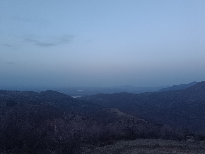 The image displays a panoramic view of a mountainous landscape under low light conditions, identified as being in Oreshets, Bulgaria. The upper third of the frame is occupied by the sky, which exhibits a gradient from a pale blue at the top to a darker, desaturated blue-grey closer to the horizon, with a solitary, wispy cloud present in the upper left. The immediate foreground consists of dark, sparse ground cover, including what appears to be dry grass and leafless shrubs or small trees. Multiple layers of hills and mountains, densely covered in dark, indistinct foliage, recede into the background, becoming progressively hazier due to atmospheric perspective. In the mid-distance, within a valley formation, at least two separate bodies of water are visible as lighter, reflective patches. No individuals, animals, or man-made structures are discernible within the depicted scene. The overall lighting and muted color palette are consistent with twilight conditions.
