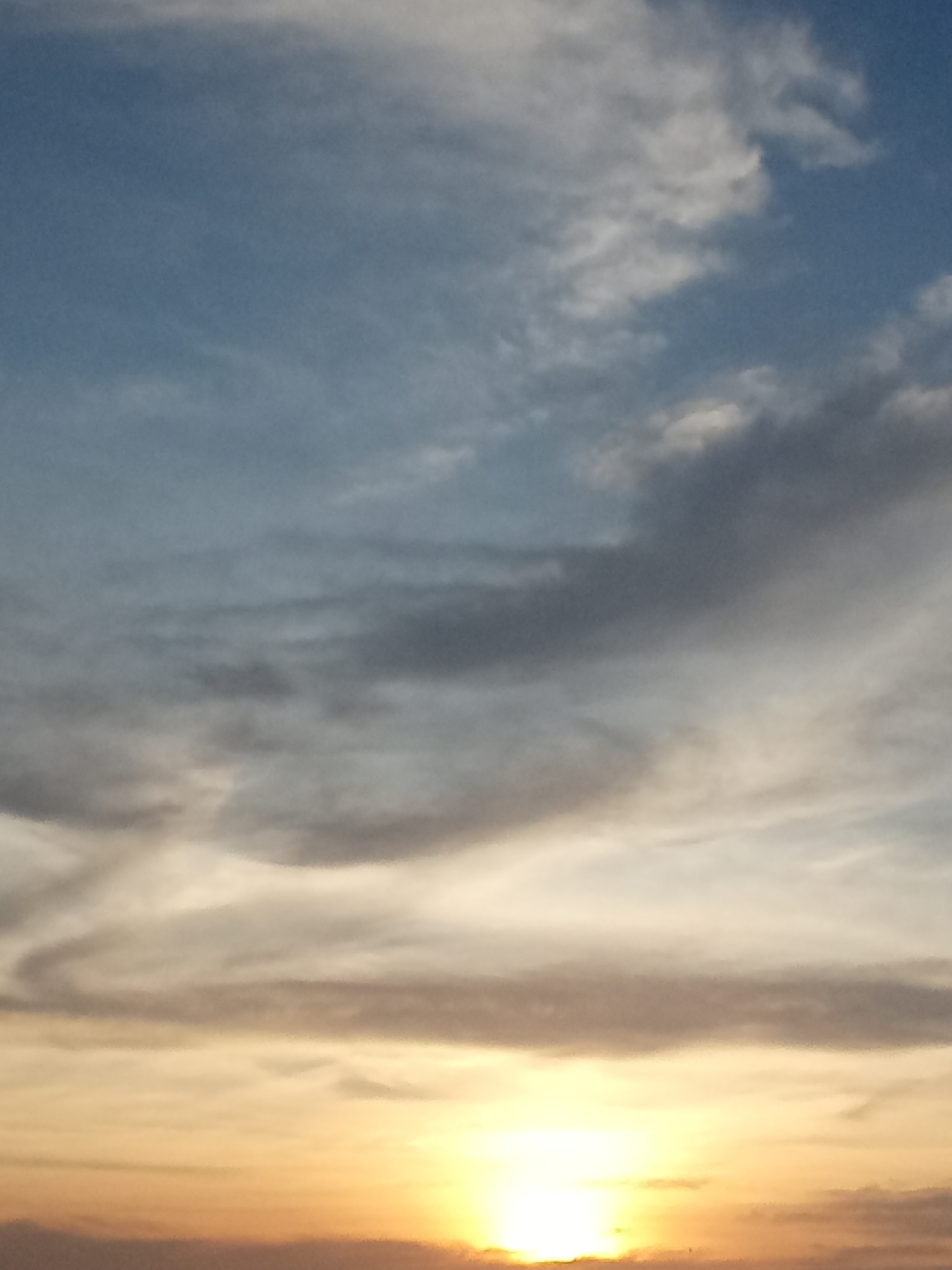 The image depicts a sunset sky in Juan Griego, Venezuela. The lower portion of the sky exhibits shades of orange and yellow, with the sun prominently visible on the horizon. Above this, bands of grey and white clouds stretch horizontally across the frame. The upper portion of the sky features a mix of light blue and scattered white clouds. There are no people or distinct objects visible beyond the atmospheric elements.