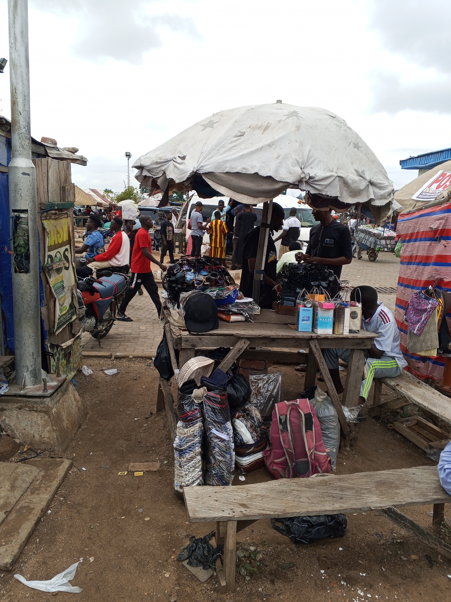 The image shows a bustling marketplace in Lokoja, Nigeria. A weathered wooden table sits in the foreground, laden with goods for sale, including sunglasses and packaged clothing. A large, star-patterned umbrella provides shade.
Several individuals are present: a man in a black shirt appears to be tending to the merchandise, while another sits on a nearby bench. People are walking and riding a motorcycle in the background, indicating a busy street.
The ground is bare earth. Other structures, including buildings and additional market stalls, are visible in the background, as well as a concrete lamp pole and a poster.
