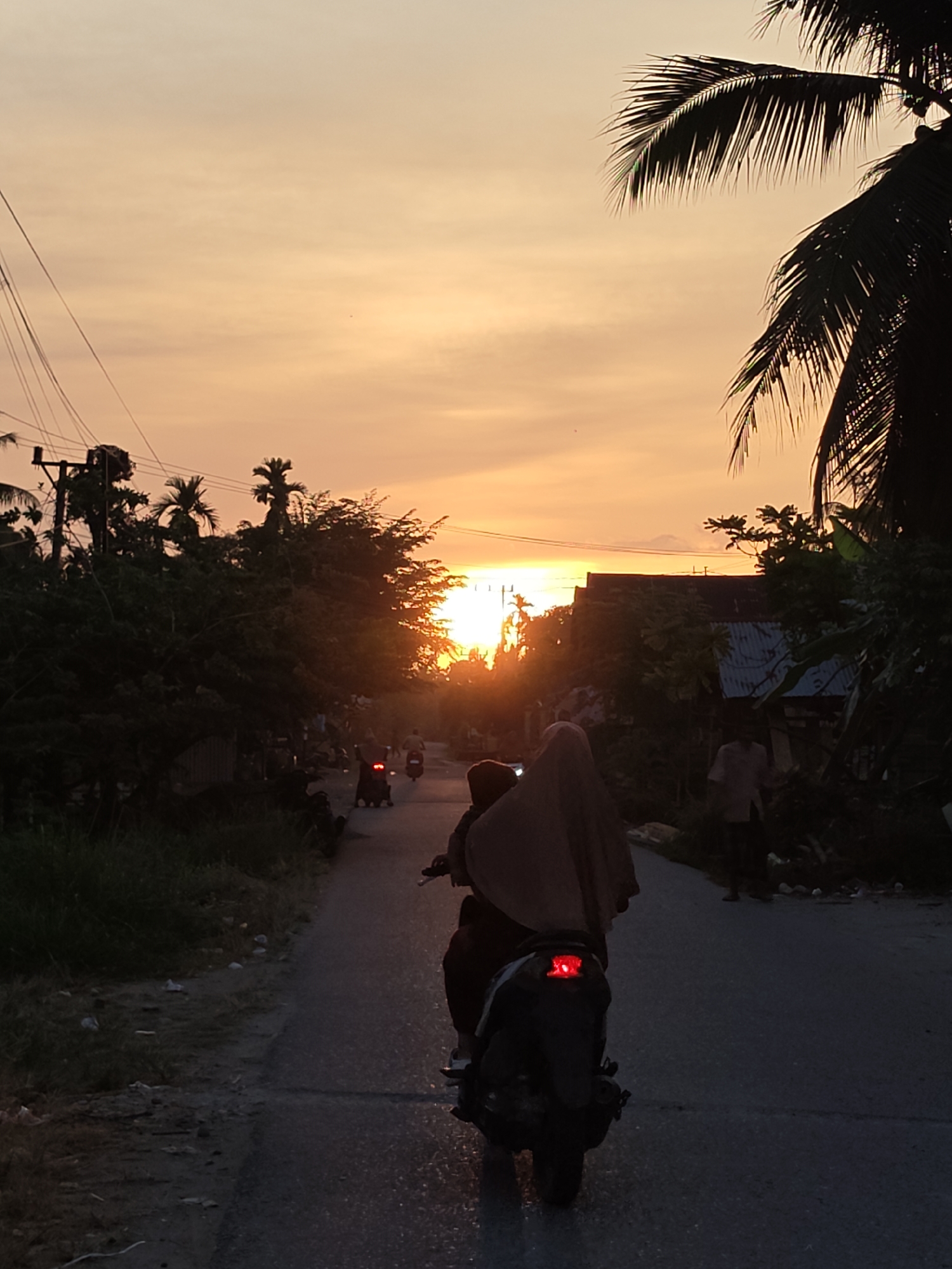 This is a photograph of a rural road in Lhokkulam, Indonesia, during a sunset. The sky is filled with warm orange and yellow hues, with a few wispy clouds. In the foreground, a motorcycle carrying two people, one wearing a hijab, travels away from the viewer. The motorcycle's taillight is illuminated red. To the sides of the road, dense foliage and palm trees are silhouetted against the bright sky. A utility pole with visible wires is also present on the left side of the frame. In the distance, other motorcycles can be seen, and several simple buildings with corrugated metal roofs are partially visible amidst the trees. The road itself appears to be paved, with some gravel and debris on the edges. The overall impression is one of a serene evening in a tropical setting.