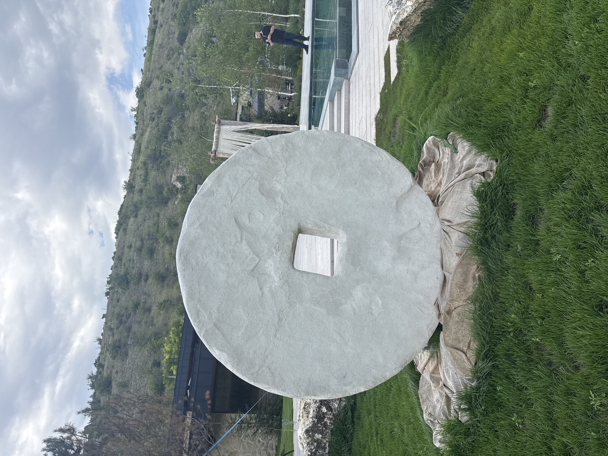 An outdoor scene captures a large, light-colored stone disc sculpture with a square hole in the center, resting on a beige cloth on a green lawn. Behind the sculpture, a swimming pool with a blue surface is visible, bordered by stone paving and steps. A man wearing a black t-shirt and pants stands on the pool's edge, holding a mobile phone. Further back, a building with dark roofing and stone walls is partially visible amidst trees. The background features a grassy hillside dotted with trees and shrubs under a cloudy sky.