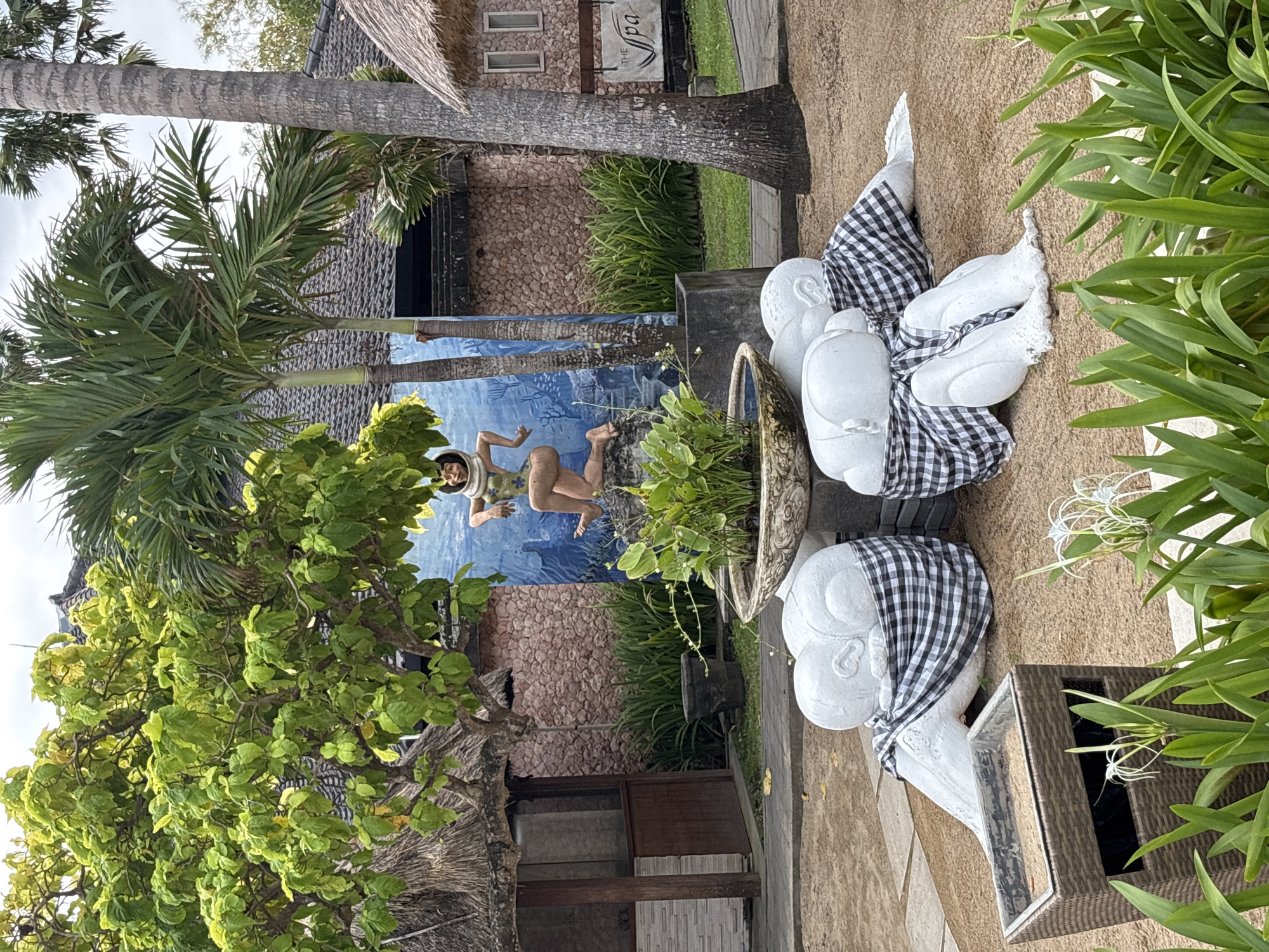 The image depicts an outdoor scene in Denpasar, Indonesia, likely a resort or spa, given the signage visible in the background. In the foreground, there are four white, abstract sculptures of figures with heads tucked into their laps, each partially draped in black and white checkered fabric. These sculptures are arranged on a sandy surface. Adjacent to the sculptures is a brown wicker trash receptacle. Further back, a decorative planter holds green foliage. Behind the planter, a mural on a blue background depicts a figure in a diving suit or similar attire, in a crouched position. To the left of the mural, there is a thatched-roof structure. The background includes a building with a stone facade and a tiled roof, palm trees, and lush green vegetation. A sign that reads "The Spa" is visible on the building to the right. The sky is overcast, suggesting it may be daytime.