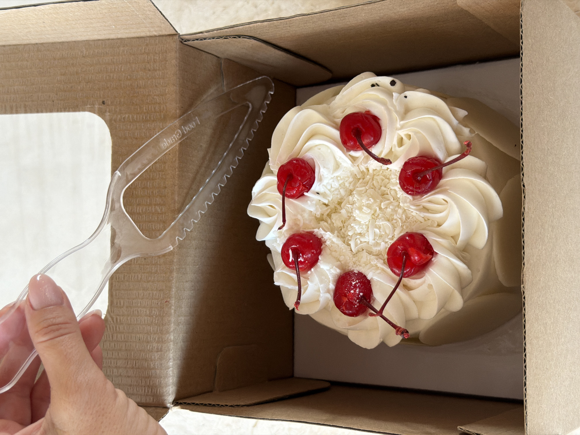 A hand holding a clear plastic cake server is above a small, round white cake decorated with swirls of white frosting, shredded white chocolate, and maraschino cherries in a brown cardboard box. The cake is positioned in the center of the box. The background is a light-colored surface.