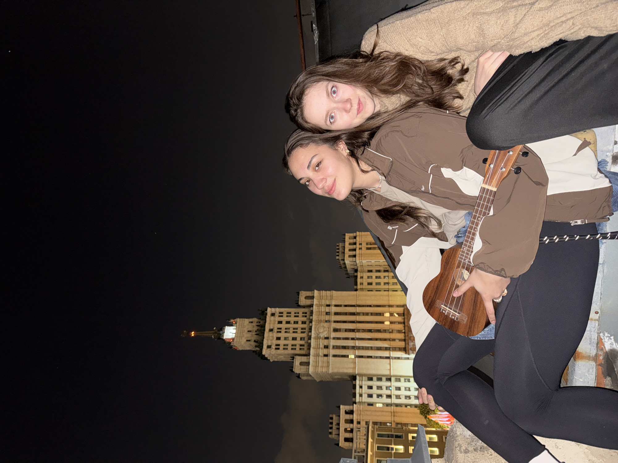 Two young women are posing for a photograph on a rooftop at night in Moscow, Russia. The first woman, in the lower part of the image, has dark hair and is wearing a brown and white jacket, black leggings, and holding a ukulele. The second woman, in the upper part of the image, has brown hair and is wearing a tan sweater and black leggings. Both women are looking at the camera and smiling. The Kotelnicheskaya Embankment Building, one of the Seven Sisters skyscrapers, is visible in the background.
