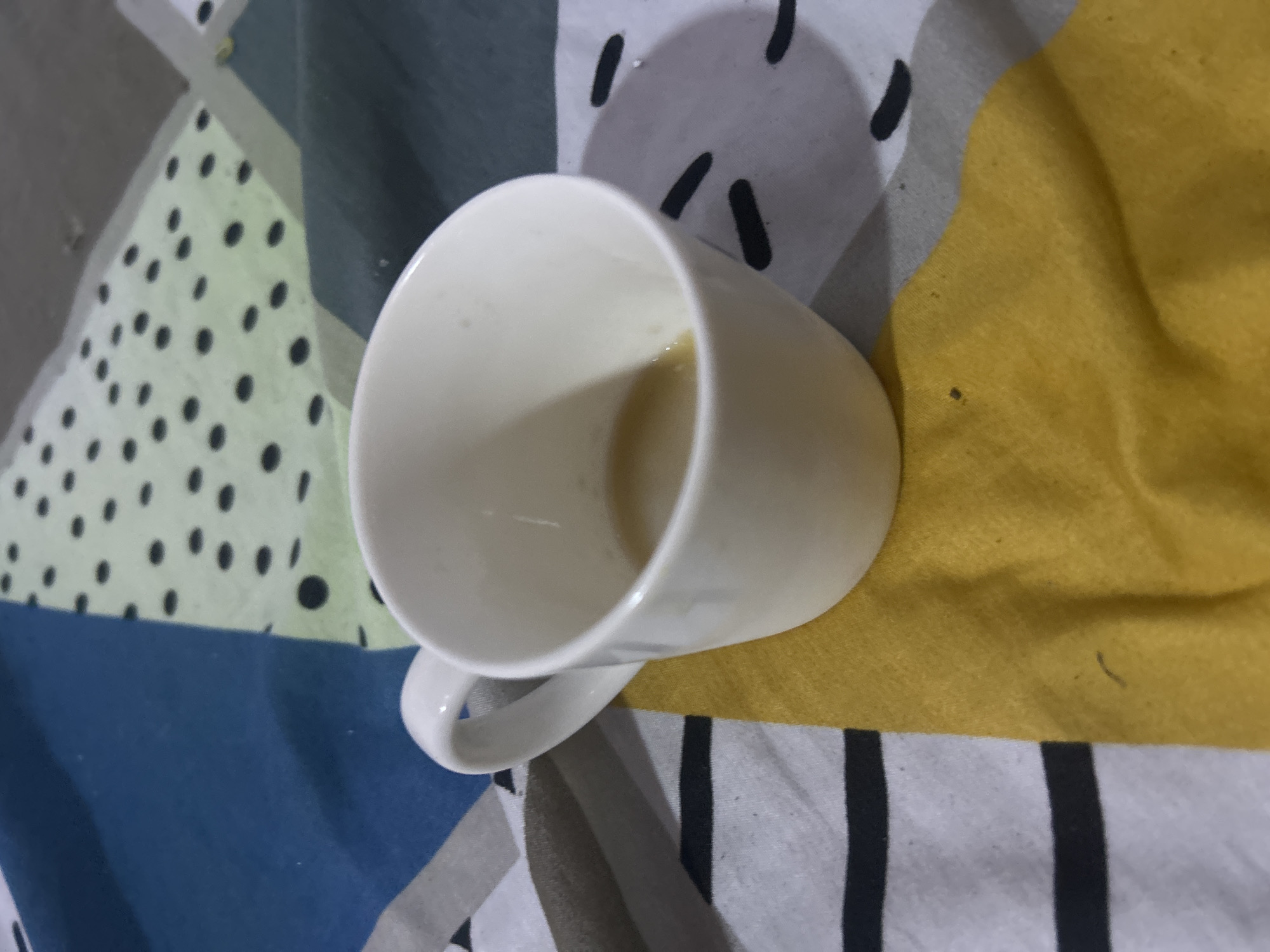 The image shows a white mug with a handle, positioned on a patterned fabric surface. The fabric has geometric patterns in shades of blue, green, yellow, gray, and white with black stripes and dots. There are residual liquid stains inside the mug. The scene's location is not discernible from the image, so I can't provide a location in San Juan de Los Morros, Venezuela.