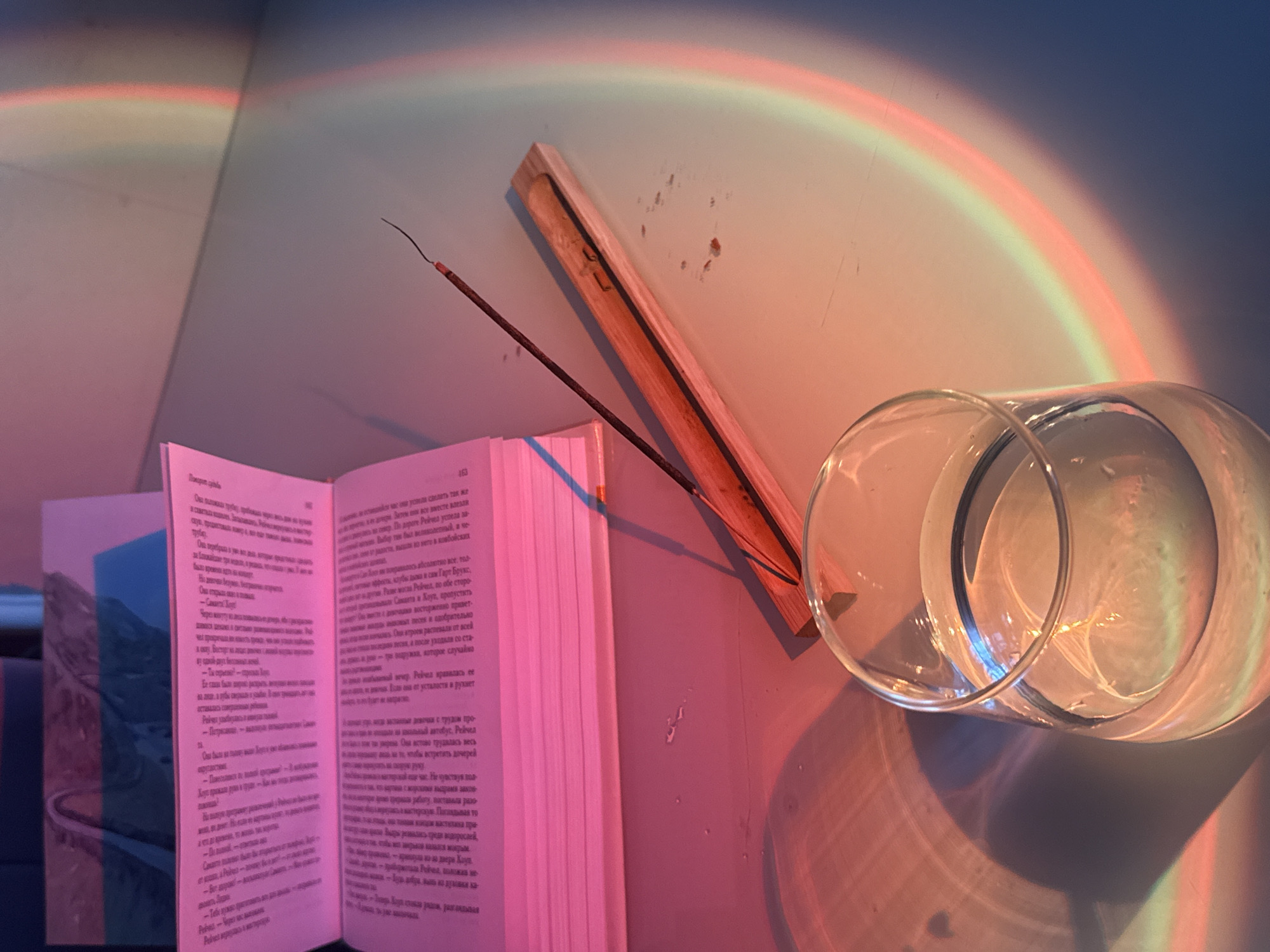 Here's a matter-of-fact description of the image:
The image presents a still life composition with several elements arranged on a surface, likely a table or shelf. An open book with text visible in a non-English language dominates the left side of the frame. A wooden holder rests near the book, containing a slender incense stick. To the right, a clear glass filled with liquid (presumably water) lies horizontally. The scene is bathed in a soft, colorful light, predominantly pink and orange, casting reflections and shadows that suggest an artificial light source or a filter effect. A light pattern reminiscent of a rainbow is cast on the wall. The setting appears to be an interior space with light.