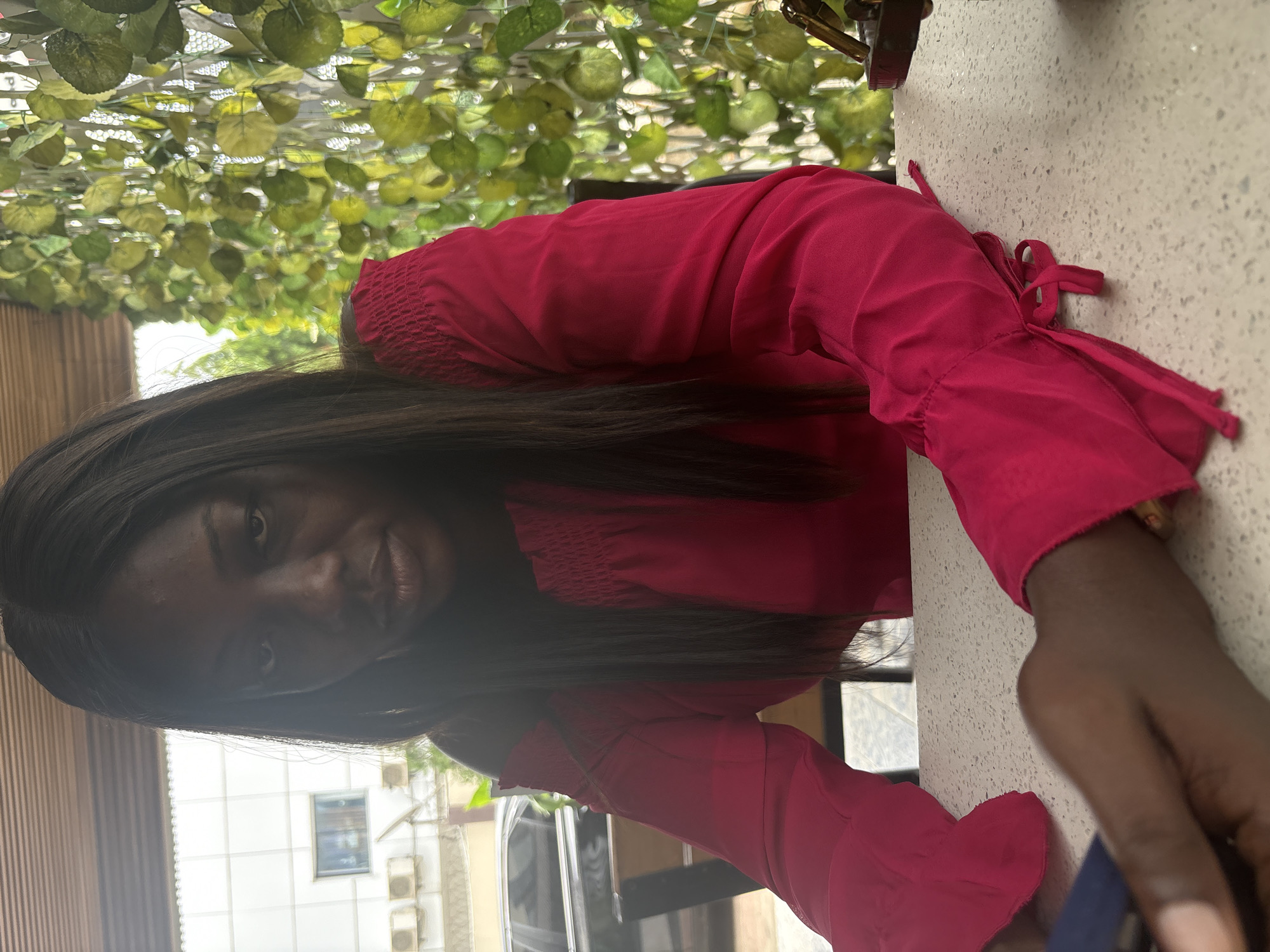 A woman with dark skin and long, dark hair is wearing a bright pink top with ruffled sleeves. She appears to be sitting at a table, as indicated by a textured, light-colored surface in the foreground. Overhead, there is an artificial covering of green leaves, resembling ivy or foliage. A building can be seen in the background with window AC units. Based on the architectural style of the building and the vegetation in the background, the location is consistent with an urban setting in Abuja, Nigeria.