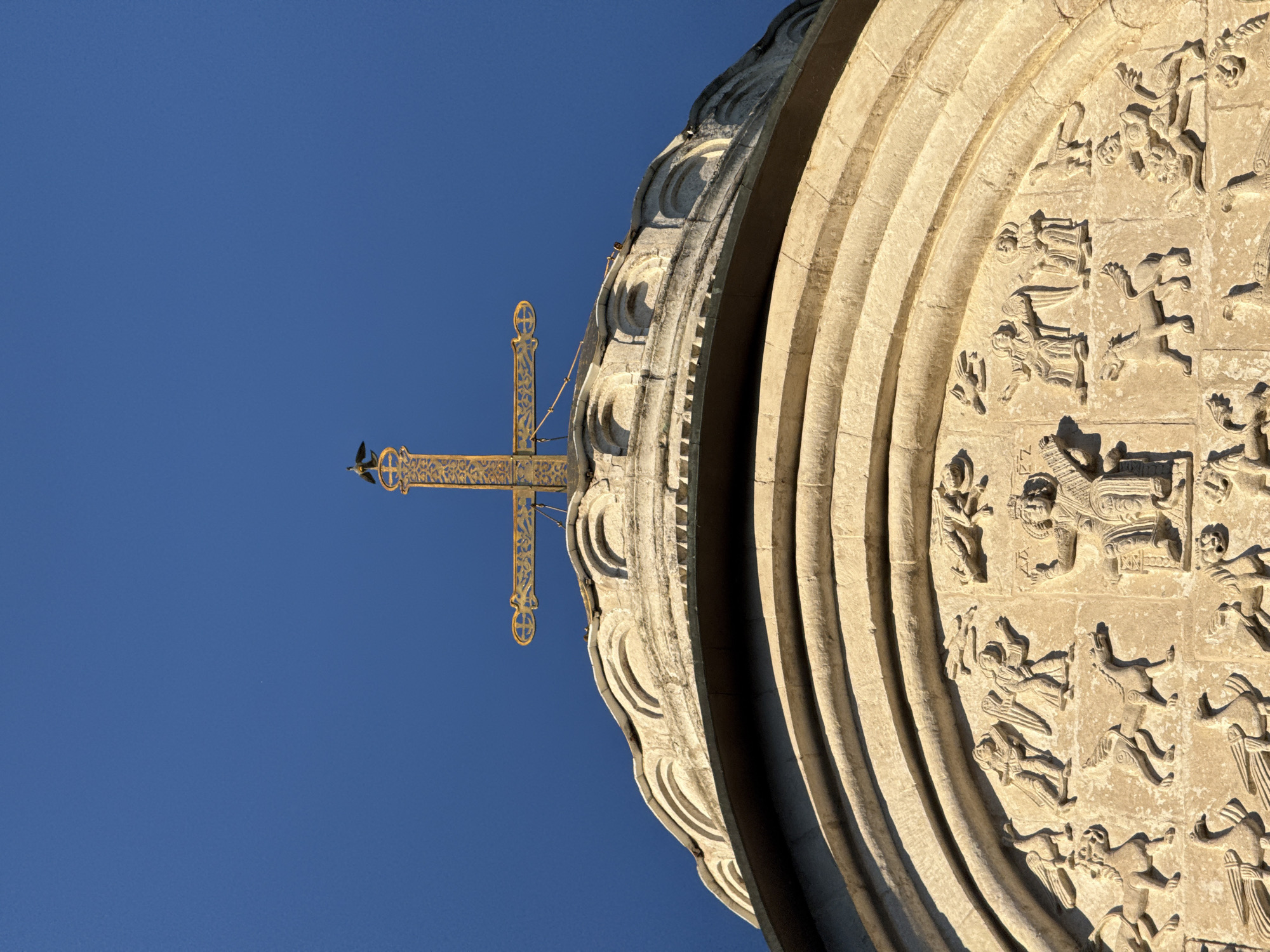 The image shows a low-angle view of a golden cross topped with a bird on a dome of the Dormition Cathedral in Vladimir, Russia. The cross is ornate, with decorative elements on its arms. The dome is made of white stone and has a series of arched carvings below the apex. The background is a clear, deep blue sky. The lower portion of the image reveals detailed stone carvings depicting figures and mythical creatures, including winged horses, birds, and human-like figures in narrative scenes.