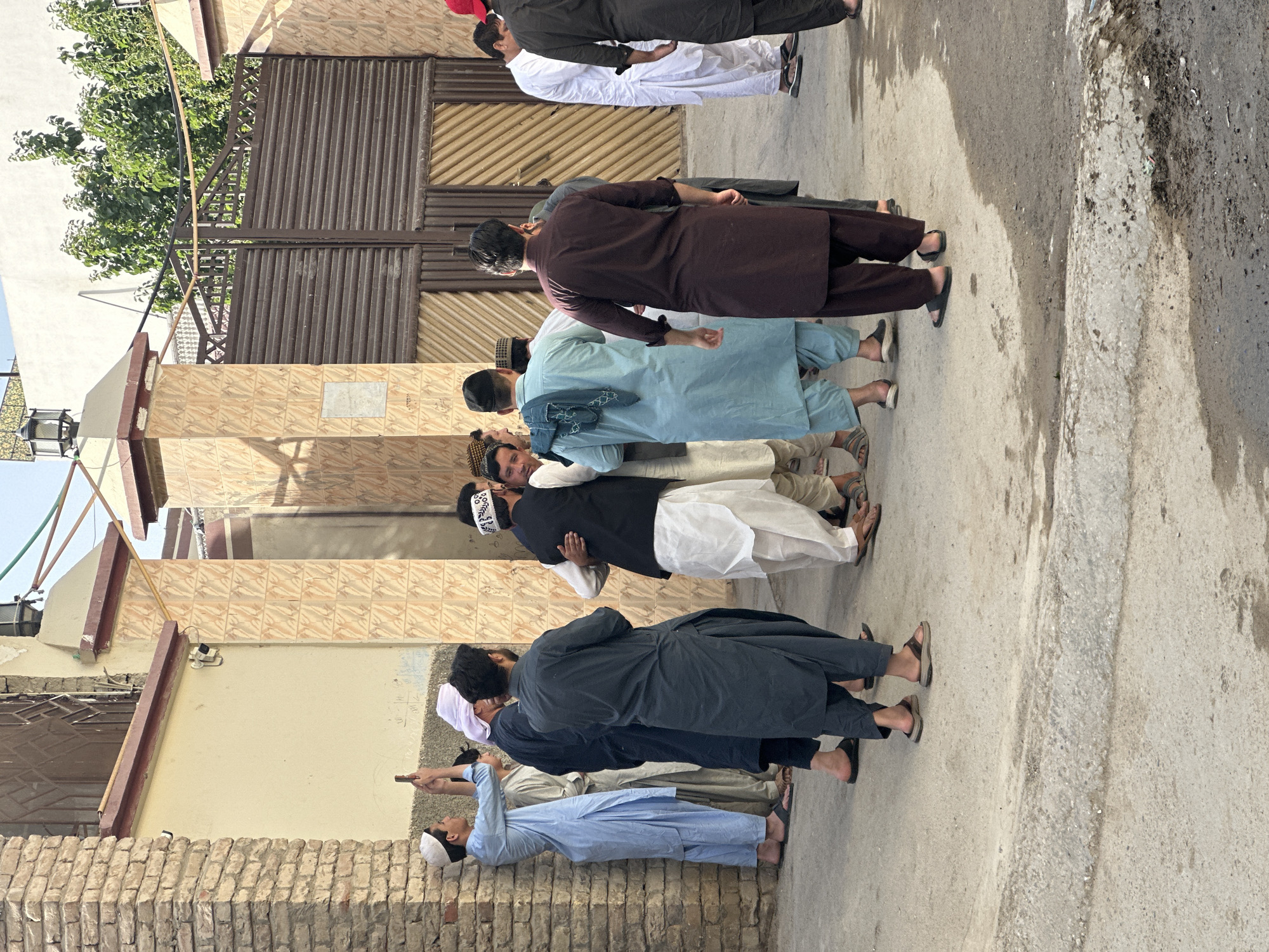 A group of men stand closely together on a street in Quetta City Tehsil, Pakistan. Most of the men are wearing traditional Pakistani clothing, including shalwar kameez in varying shades of blue, brown, black, and white. Some wear white or patterned skullcaps. They are on a paved street with buildings and a metal gate behind them.