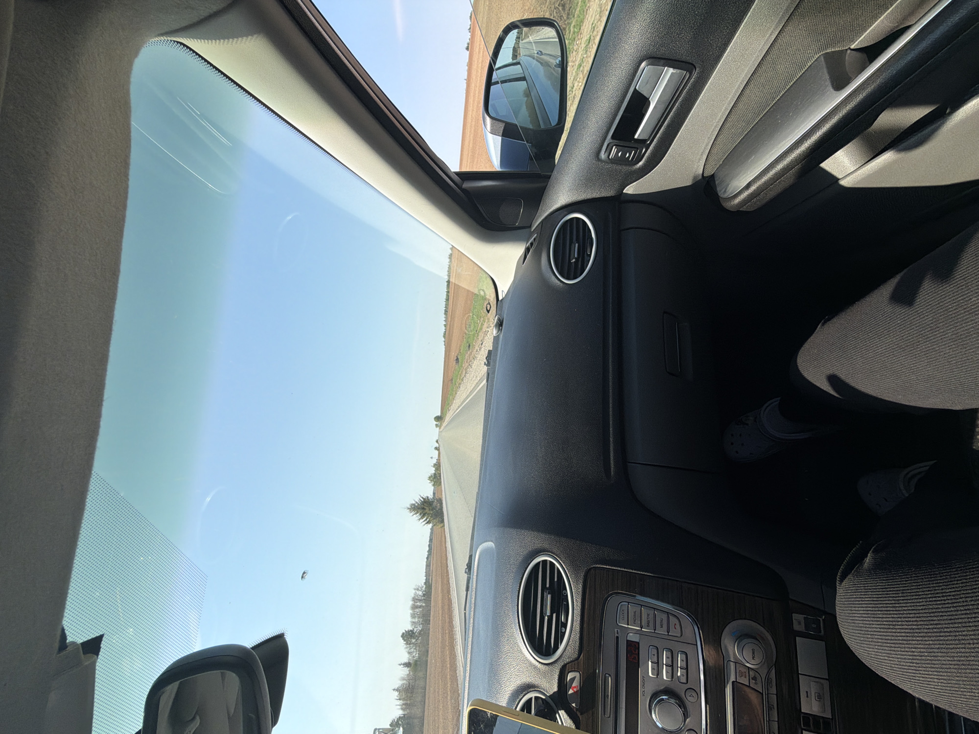 The image shows the interior of a car driving on a road near Перевозное, Russia. The dashboard and steering wheel are visible. The road is surrounded by fields, and the sky is clear. The driver is wearing black pants and Crocs. The image is taken from inside the vehicle, looking out the windshield. A phone is attached to the dashboard.