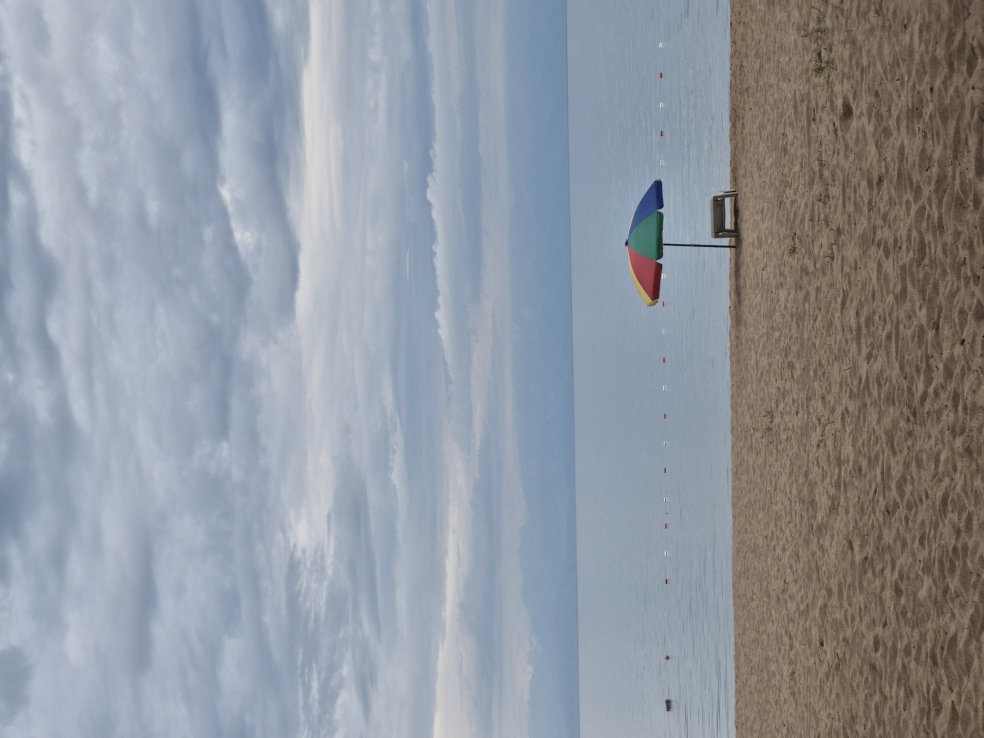 The image depicts a tranquil beach scene in Bulan-Segettu, Kyrgyzstan. The foreground is dominated by coarse, light brown sand, textured with ripples and small indentations, suggesting a natural, undisturbed state. A few sparse blades of dry grass are visible on the sand. In the midground, a calm, expansive body of light blue water stretches towards a hazy horizon, where faint, muted mountain ranges are discernible. A line of small, alternating red and white buoys marks a boundary in the water. A solitary, multi-colored beach umbrella, featuring segments of red, yellow, green, and blue, stands on the sand near the water's edge. Beneath the umbrella is a simple, light-colored lounge chair. The sky above is overcast with a dense layer of grey clouds, creating a subdued, diffused light. The overall atmosphere is one of quietude and natural beauty.