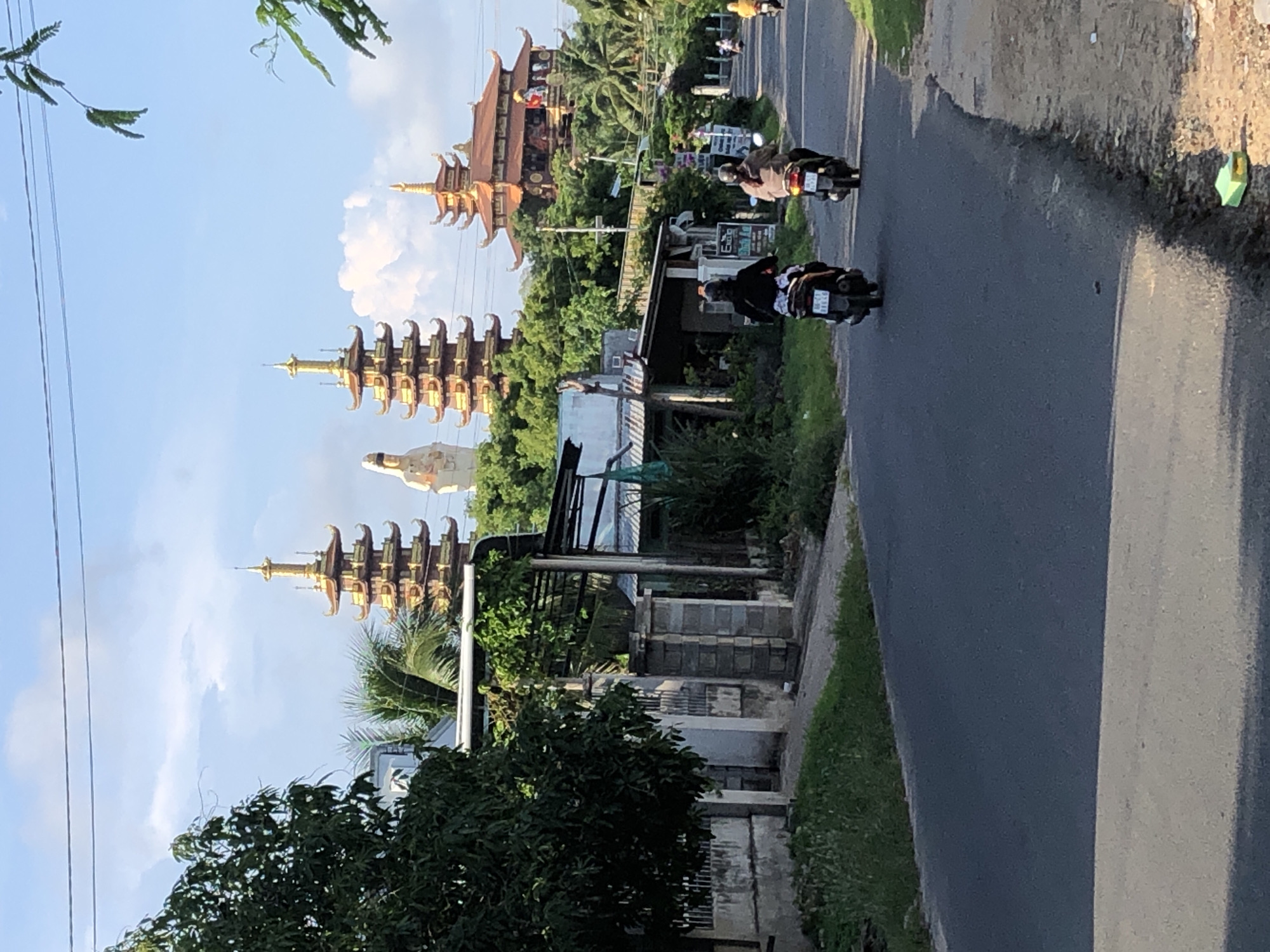 A sunny day in Mui Ne, Vietnam, features two pagodas with golden spires and ornate red roofs. A large white statue stands between them. In the foreground, a paved road is used by several motorbikes. Two people on motorbikes are traveling away from the camera, with one on the left and another on the right, both wearing helmets and casual clothing. A third motorbike with a person wearing a yellow shirt is visible further down the road. Along the side of the road, there are houses, trees, and greenery, including a large tree on the left and palm trees on the right. The sky is blue with some clouds, and telephone wires are visible overhead. The scene captures a slice of daily life in Mui Ne, with its distinctive architecture and modes of transportation.