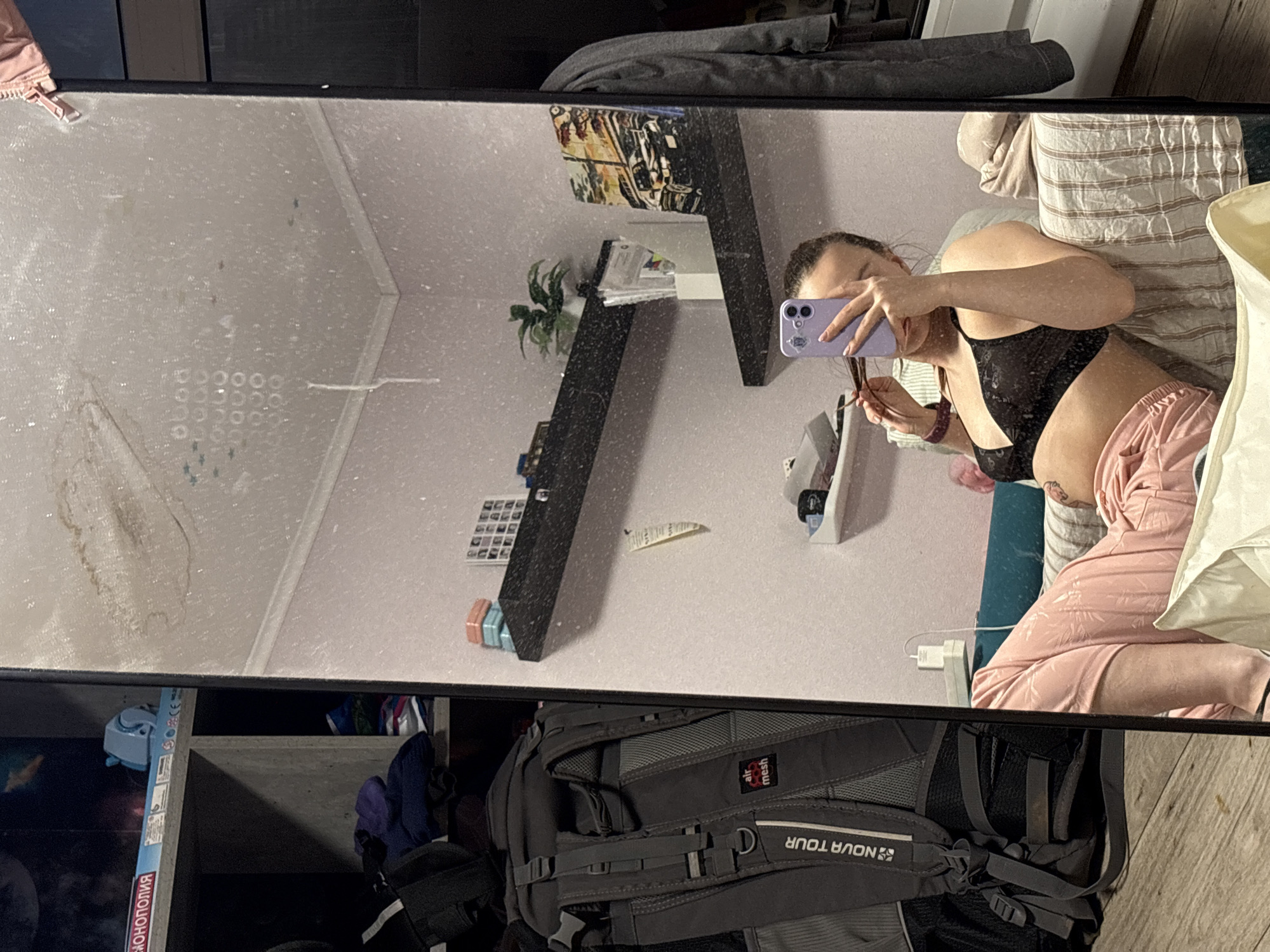 The image captures a mirror selfie taken indoors in Прикамский, Russia. A woman is visible in the reflection of a large, rectangular mirror with a black frame. She is wearing a black bra or sports bra and light pink patterned shorts or pants. She holds a purple smartphone with multiple camera lenses to take the photo, partially obscuring her face. A watch or bracelet is on her left wrist, and a tattoo is visible on her left arm.
The mirror surface is noticeably dirty, covered in numerous smudges, dust, and watermarks.
The reflected background shows a room with light-colored walls and a dark, multi-tiered shelf unit. The shelves hold various items, including a green potted plant, books, and other decorative objects. A light-colored striped pillow or blanket is seen next to the woman in the reflection.
In the foreground of the actual image, below the mirror, a grey backpack with the text "NOVA TOUR" and "air mesh" is visible. Adjacent to it are dark storage shelves containing clothes and other items. A board game box, partially seen, displays the Russian word "МОНОПОЛИЯ" (Monopoly). The lighting suggests an indoor setting, likely with artificial light. The image depicts a casual, everyday moment in a personal living space.