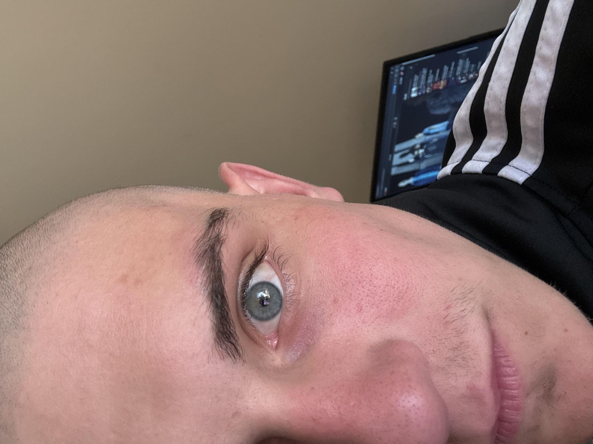 A close-up, horizontally oriented image features a person with light skin, a prominent blue-grey eye, and a dark eyebrow. The person's head appears to have very short, possibly shaved hair or stubble on the side, with visible skin texture, including pores and some redness on the cheek. A small portion of the nose and lips are visible, with faint stubble on the upper lip. The person is wearing a black garment with three distinctive white stripes, characteristic of Adidas athletic wear.
In the background, a computer monitor is partially visible, displaying a dark graphical user interface with various panels and elements, suggesting a software application such as a video editor or a game. The eye reflects a rectangular light source, likely the monitor. The background wall is a plain, light brown/beige color. The image captures an individual engaged with a computer in an indoor setting, possibly in Varna, Bulgaria. No legible text is visible within the image itself.