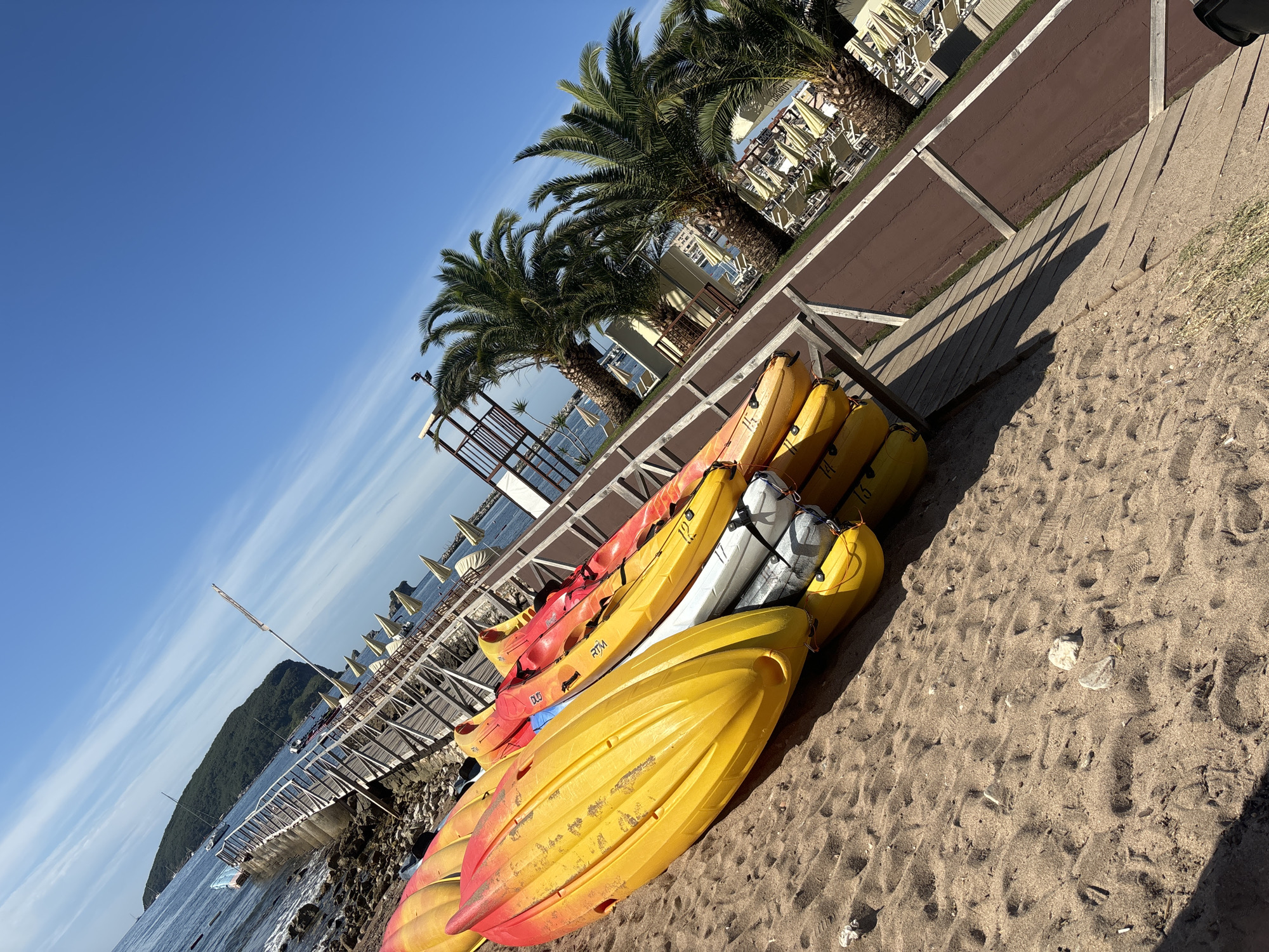 The image depicts a sunny day at a beach in Budva, Montenegro. In the foreground, a stack of brightly colored kayaks, primarily yellow and red, rests on the sandy shore. To the left, the turquoise waters of the Adriatic Sea meet a rocky coastline with a wooden pier extending into the water. Further back, palm trees line the promenade, with lounge chairs and umbrellas visible in the distance. The sky is a clear, vibrant blue with a few wispy clouds. The overall scene suggests a recreational area, likely a tourist destination, ready for water activities.