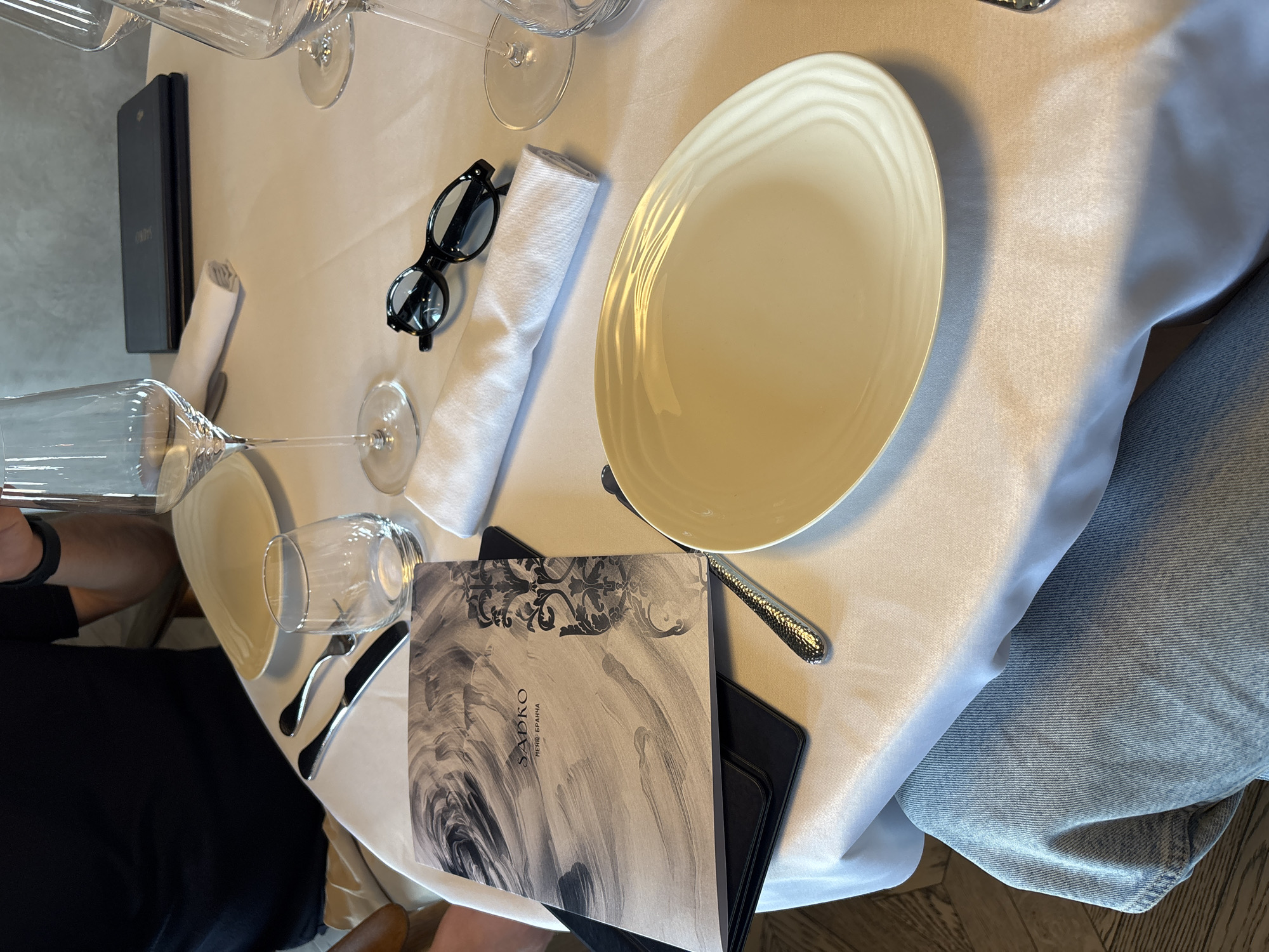 This image presents an overhead view of a neatly set table within what appears to be a restaurant, indicating preparation for a meal. The table is draped with a pristine white tablecloth, reflecting a clean and elegant dining environment.
Central to the setting is an empty, light-colored, oval-shaped plate, positioned for a diner. It is accompanied by a full set of silverware, including a fork, knife, and a spoon with a textured handle. A white rolled linen napkin lies to the left of the plate, topped by a pair of black-framed eyeglasses. Multiple empty wine glasses and a water glass are arranged around the setting, suggesting a complete beverage service.
A prominent detail is the menu positioned on the lower right side of the table. The cover displays "SADKO" in large letters, with "МЕНЮ БРАНЧА" (which translates to "BRUNCH MENU") written beneath it. The menu features an intricate design of swirling black and grey patterns. A black folder or placemat is visible directly beneath the menu. On the far left, another black booklet or small electronic device in a case is partially visible.
Two individuals are subtly present: an arm wearing a dark watch is seen on the left edge, and a leg clad in blue denim is visible under the table on the right. The "Brunch Menu" indicates that the event is taking place during the late morning or early afternoon. The Cyrillic text on the menu further places the scene within Russia, consistent with the provided location context of Krasnoyarsk. The overall scene conveys a calm and sophisticated dining experience.