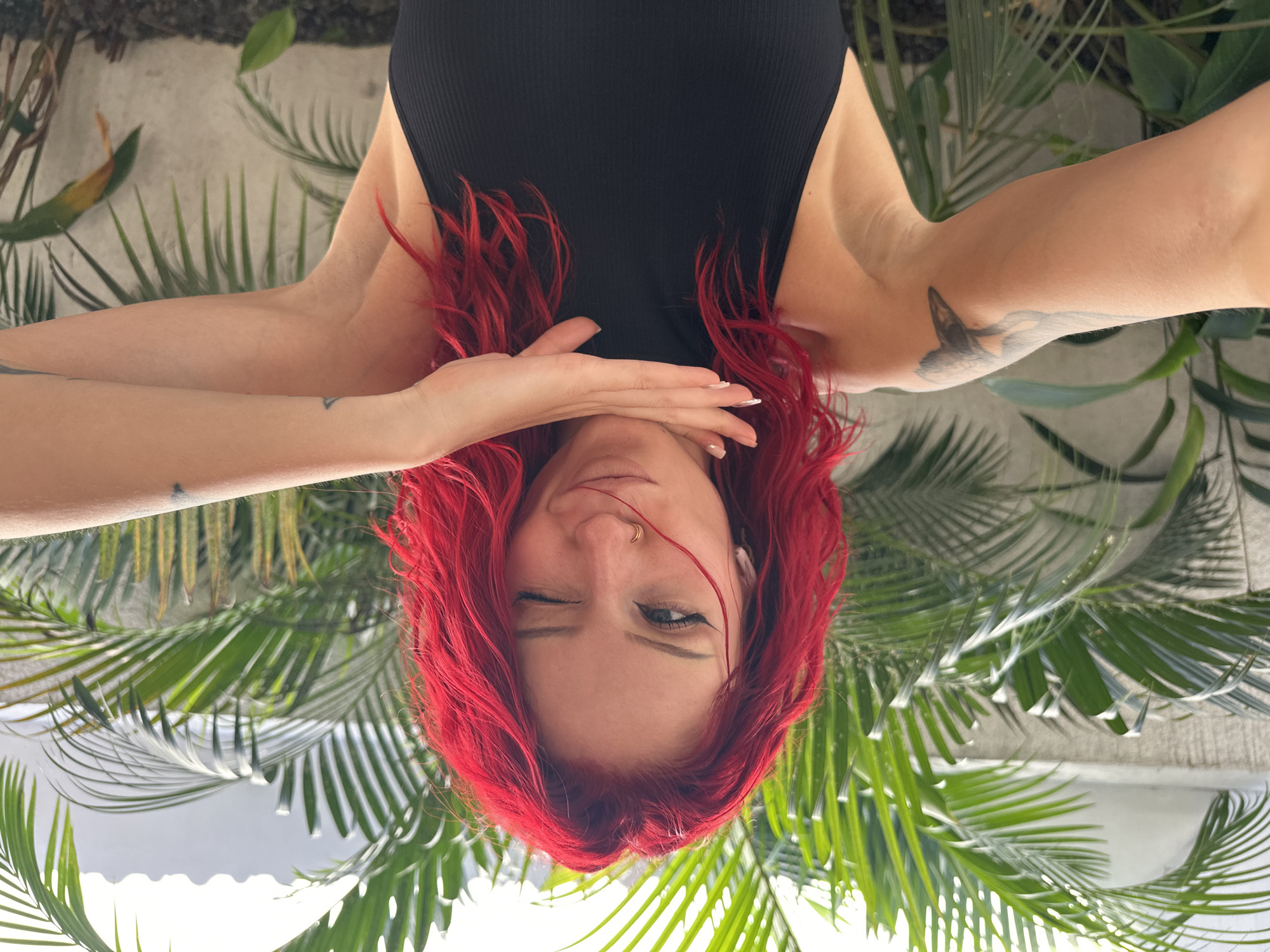 A woman with red hair and a black top is positioned upside down in front of palm trees. She is winking with her left eye, her right eye is open, and her left hand is partially covering her face. Both of her arms are outstretched on either side. She has tattoos on both arms. The background includes a wall and lush tropical foliage. The scene is outdoors in North Kuta, Indonesia.