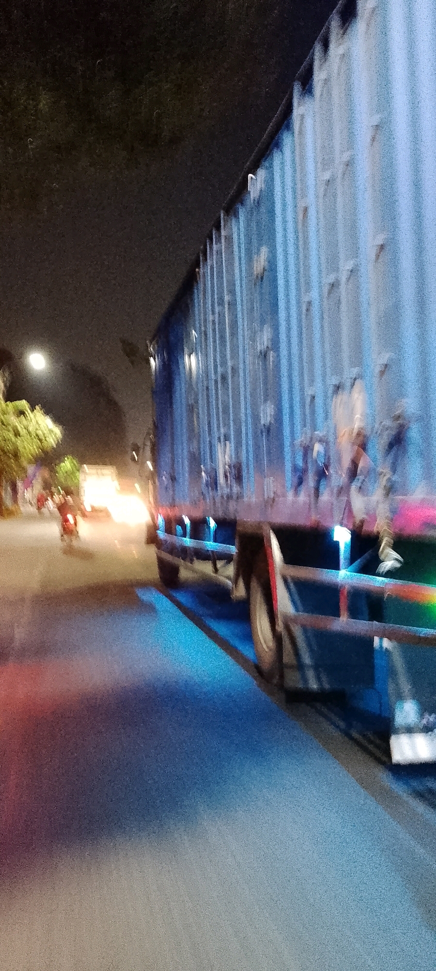 An out-of-focus, nighttime exterior shot captures a large blue truck with ribbed sides driving on a street in Bekasi, Indonesia. The truck is the dominant subject, partially obscuring the night sky which is dimly lit by artificial lights and a full moon. The street is wet, reflecting the lights from the truck and distant traffic. To the left of the truck, a motorbike carrying two individuals is visible, also in motion and slightly blurred, contributing to the sense of movement. The background shows trees and streetlights, consistent with an urban environment in Bekasi. The overall impression is one of a busy, nocturnal urban scene.