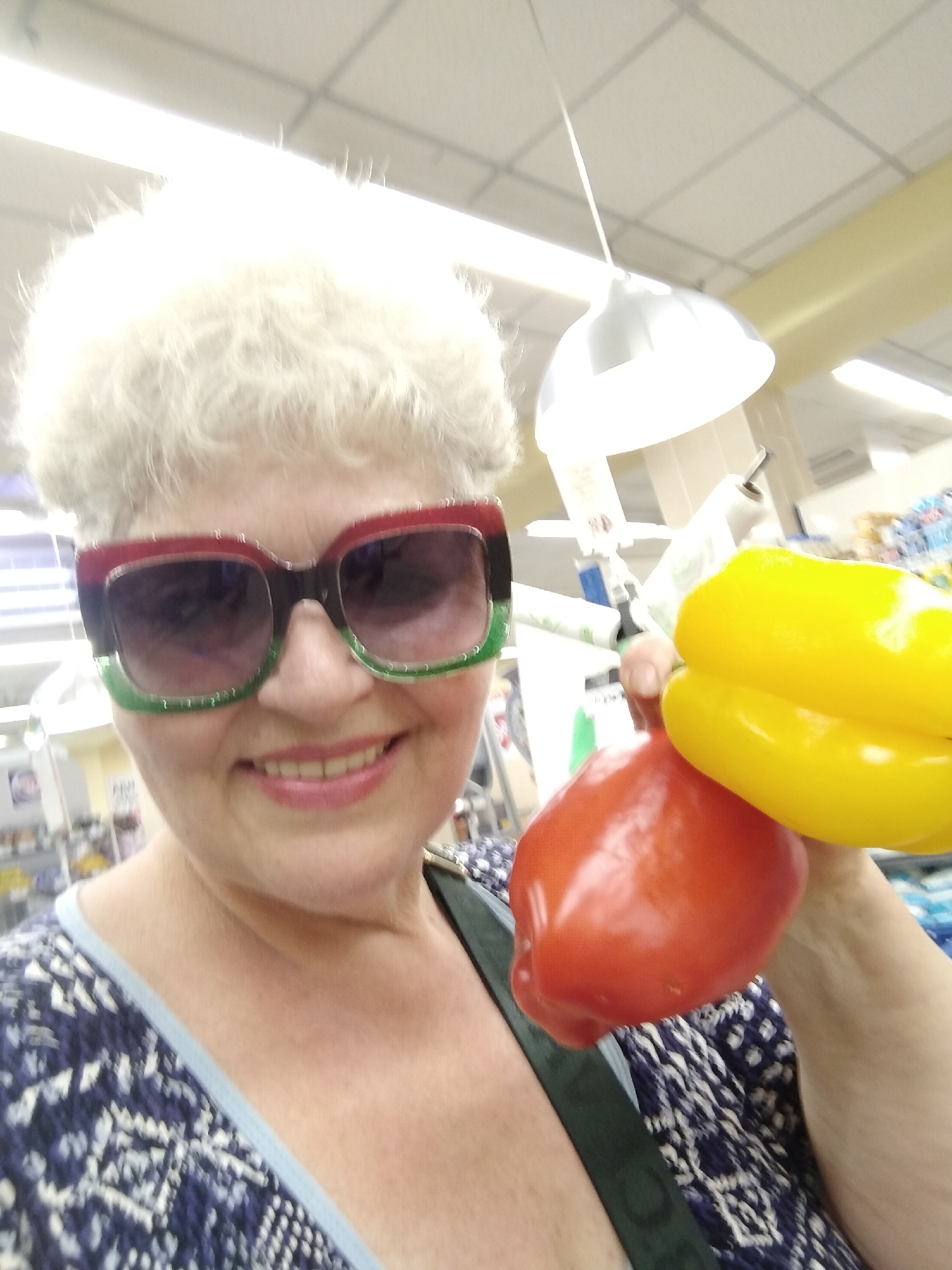 A woman with white, curly hair and large, colorful sunglasses is holding two bell peppers, one red and one yellow, in front of her face. She is wearing a blue and white patterned shirt. The background appears to be inside a store, with shelves and lighting visible. The scene is brightly lit. This image was taken in Horishni Plavni, Ukraine.