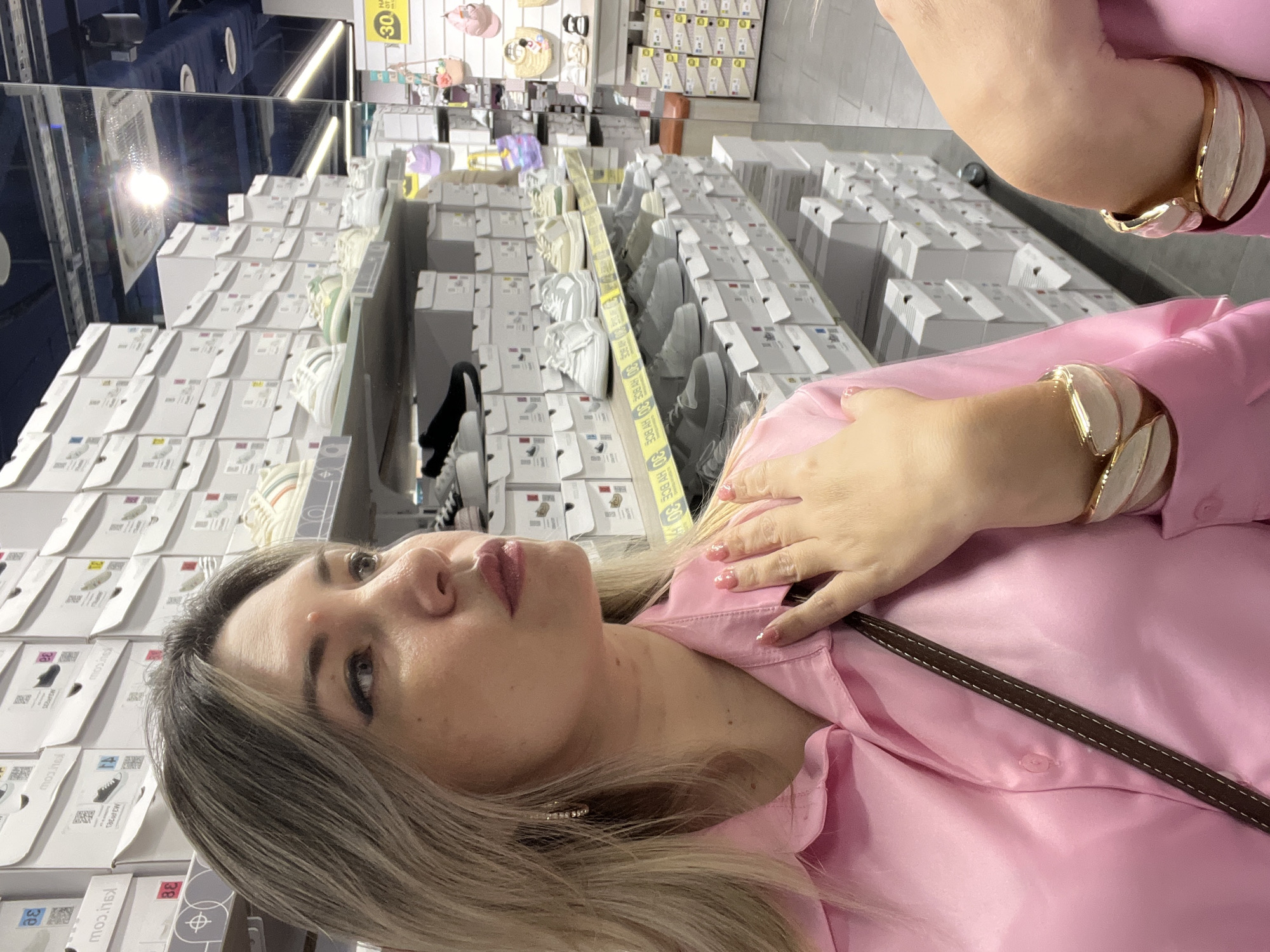 The image depicts a woman in a pink silk shirt in a shoe store. She is seen from the chest up, with her left hand resting on her chest. The background is filled with shelves of shoe boxes, indicating a retail environment. In the foreground, a portion of another person's arm with a similar pink garment and a gold bracelet is visible. The scene appears to be indoors, in a well-lit commercial space. The woman has blonde hair and is wearing hoop earrings and a bracelet. She has a small blemish on her forehead. The shoe boxes are stacked high, suggesting a large inventory. A reflection on the ceiling indicates the presence of mirrors or other reflective surfaces in the store.
