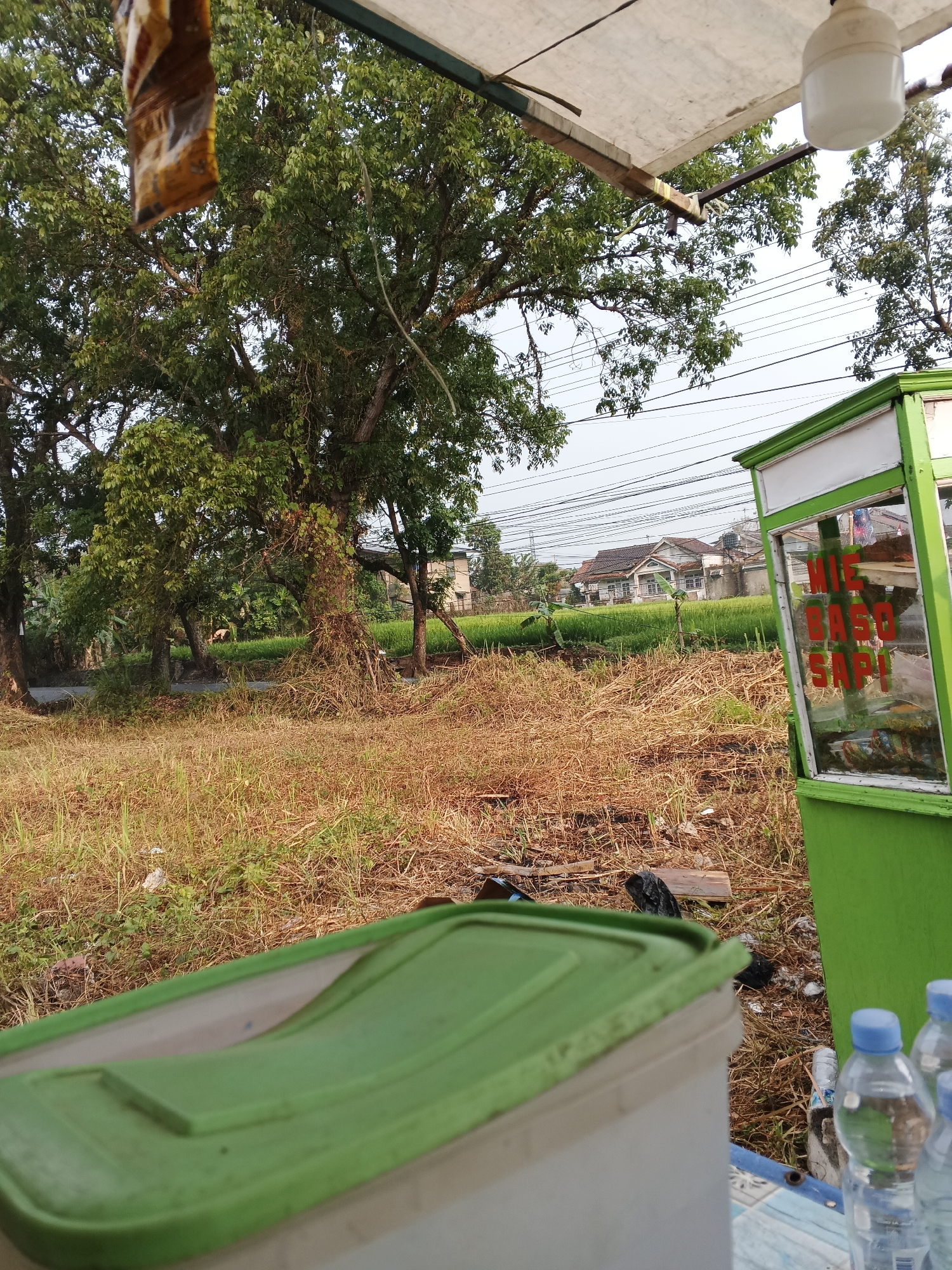 An outdoor scene in Sukabumi, Indonesia, captured from a low angle. In the foreground, a green plastic lid is visible, partially obscuring a white container. To the right, a green food cart stands, its glass display showing items and red text that reads "NIE BASO SAP!". Behind the cart, a field of dry, brown grass stretches towards a residential area with houses. Several trees with dense green foliage are scattered throughout the scene, and power lines are visible overhead. The overall impression is one of a humble, everyday setting, possibly near a roadside food stall.
