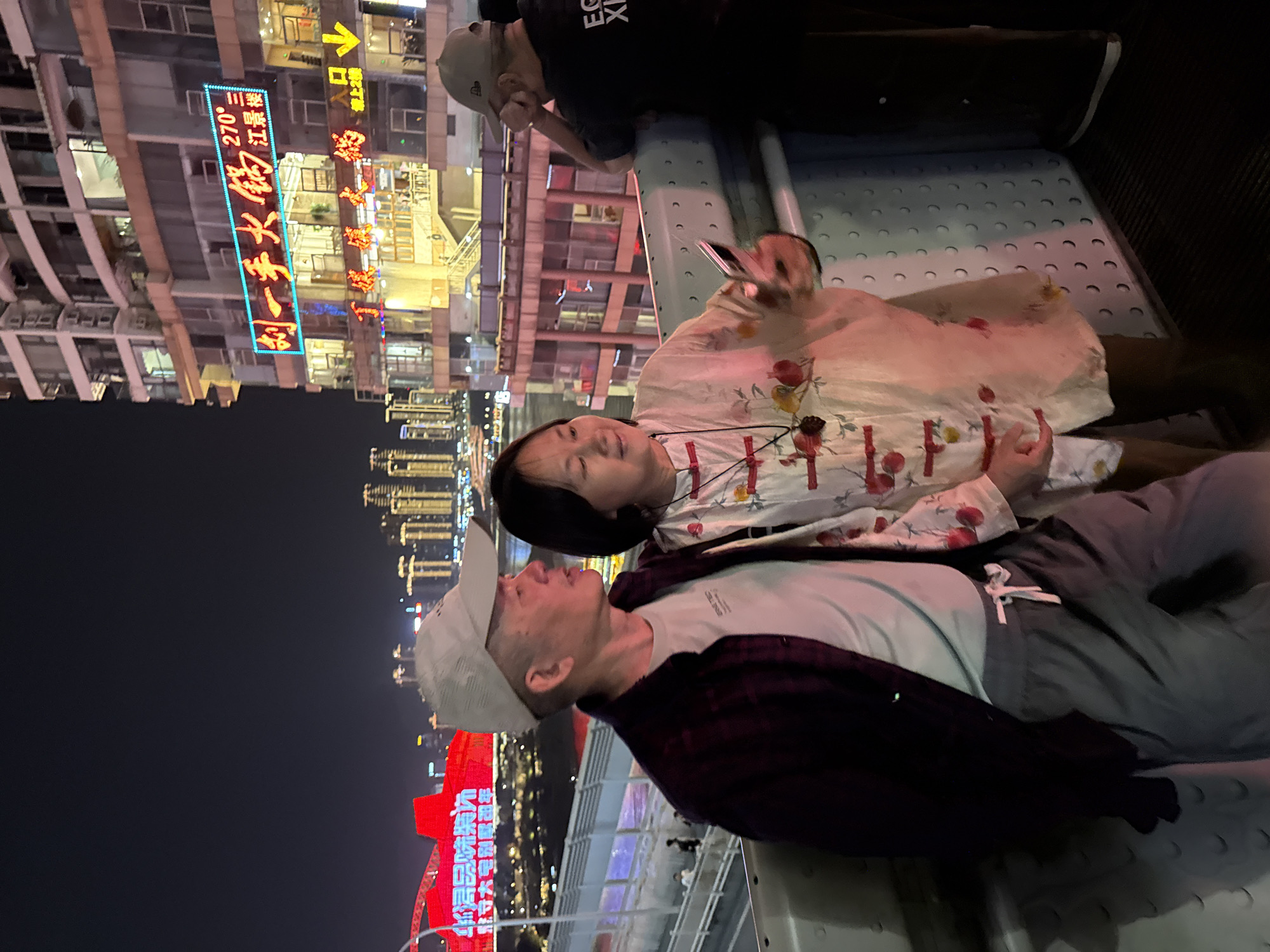 The image captures a vibrant nighttime scene in Yuzhong District, China, likely from an elevated viewpoint or walkway overlooking a cityscape. In the foreground, a man and a woman are the central subjects. The man, wearing a light baseball cap, a white t-shirt, and a dark plaid shirt, is looking towards the woman. The woman, with dark hair, is wearing a light-colored top patterned with fruits or flowers, and appears to be smiling or laughing as she looks towards the man, holding a mobile phone in her hand.
Behind them, another person, partially visible and wearing a dark shirt with "EC XI" text and a light cap, leans on a railing, engrossed in their own mobile phone. The setting is illuminated by numerous artificial lights from the surrounding high-rise buildings and prominent neon signs.
Notable details include a striking blue neon sign on a building in the background that reads "刘一手火锅" (Liu Yishou Hotpot), along with "270°" and "三楼" (Third floor) and "江畔" (Riverside), suggesting a panoramic hotpot restaurant location. Other visible Chinese text includes "华得品味裝", "駐守大宅别摩司年", "BRNE TIED", "入口" (Entrance) with an arrow, and "دور". The overall atmosphere is lively and urban, characteristic of a bustling city at night.
