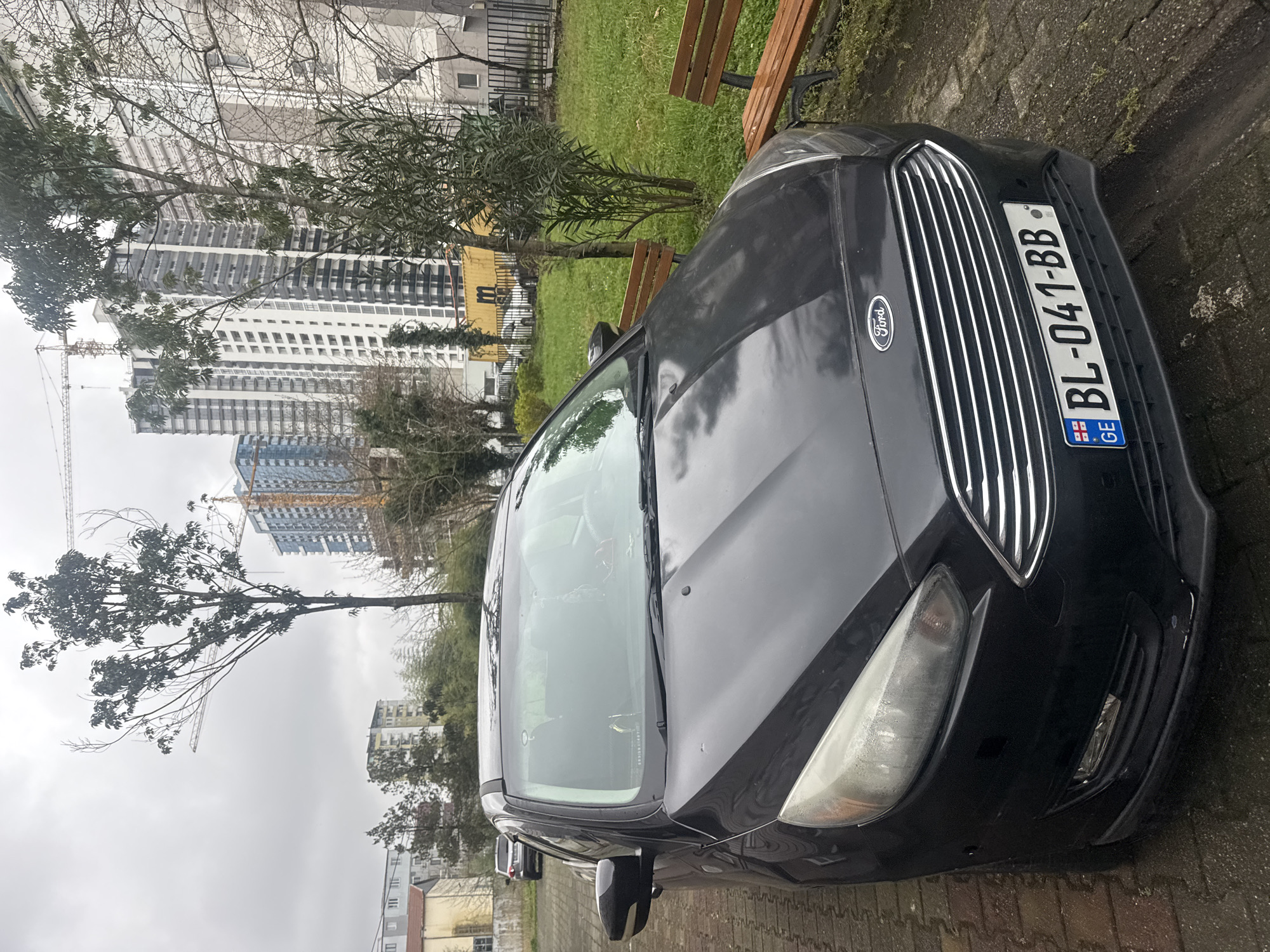 The image depicts an urban street scene in Batumi, Georgia, under an overcast sky. A dark gray/black Ford sedan, license plate GE BL-041-BB, is parked on a wet, interlocking paver surface in the foreground, angled slightly left. The vehicle's front passenger headlight shows internal condensation or haziness, and minor scuff marks are visible on the lower front bumper. Adjacent to the vehicle's right side is a grassy area containing two unoccupied wooden park benches with black metal frames, along with several trees and dense bushes. In the background, multiple high-rise residential buildings are visible, with tower cranes indicating ongoing construction for some. Older, lower-rise apartment blocks are also present on the left side of the frame. No individuals are observed within the scene.