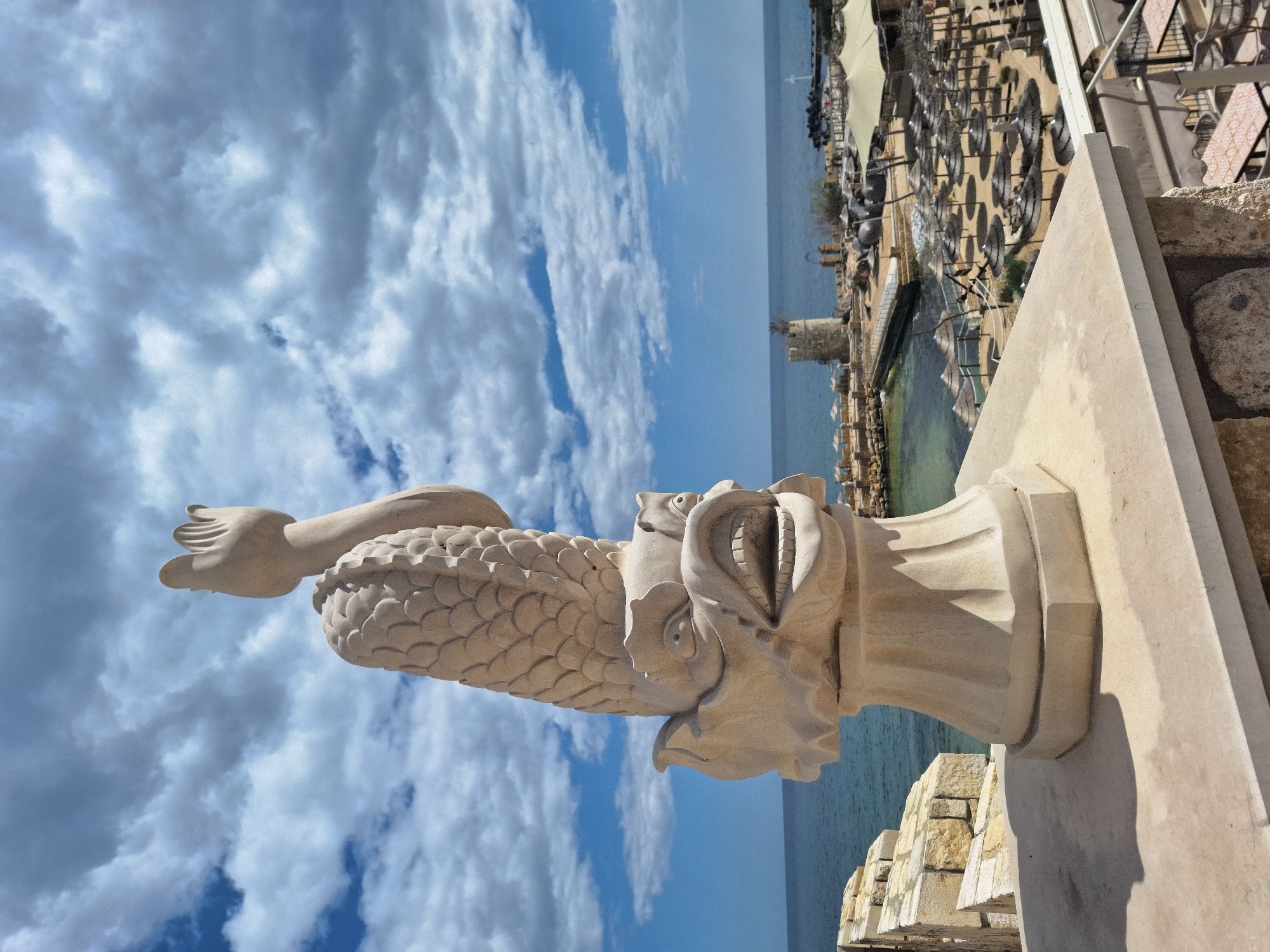 The image features a stone statue of a mythical creature with a fish-like body, a human hand, and a grotesque face. The statue appears to be part of a building's facade, overlooking a seaside view. Below the statue, there's a waterfront area with pools, outdoor seating (tables and chairs with parasols), and possibly a restaurant or cafe. In the background, there is the sea and a partly cloudy sky with white clouds against a blue background. Based on the style of architecture and the coastal setting, the location is likely the city of Varna, Bulgaria.